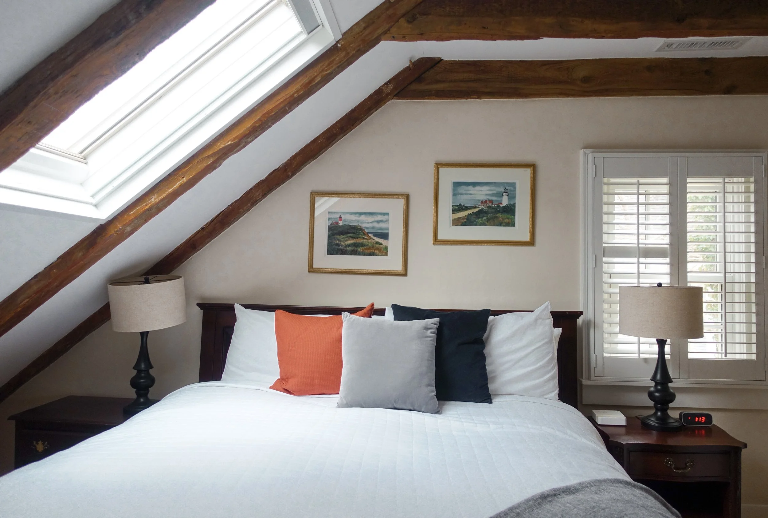 A cozy bedroom with a sloped ceiling and a skylight window, a bed with white bedding, and five pillows in orange, gray, black, and white. Two matching lamps are on dark wooden nightstands, and framed landscape paintings hang above the bed. There is a