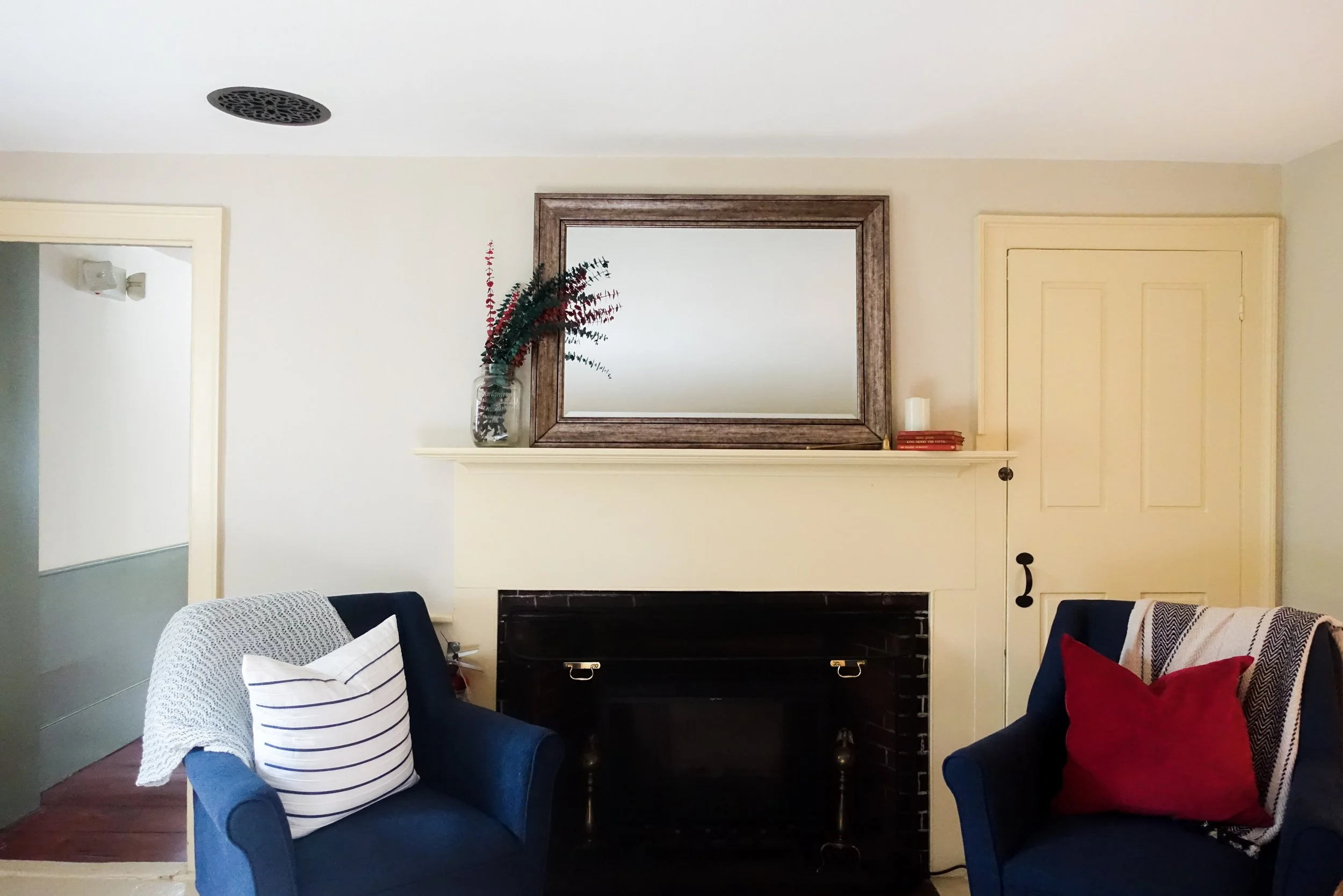 Living room with a fireplace, large mirror above, two navy armchairs with cushions, and a vase of greenery on the mantle.