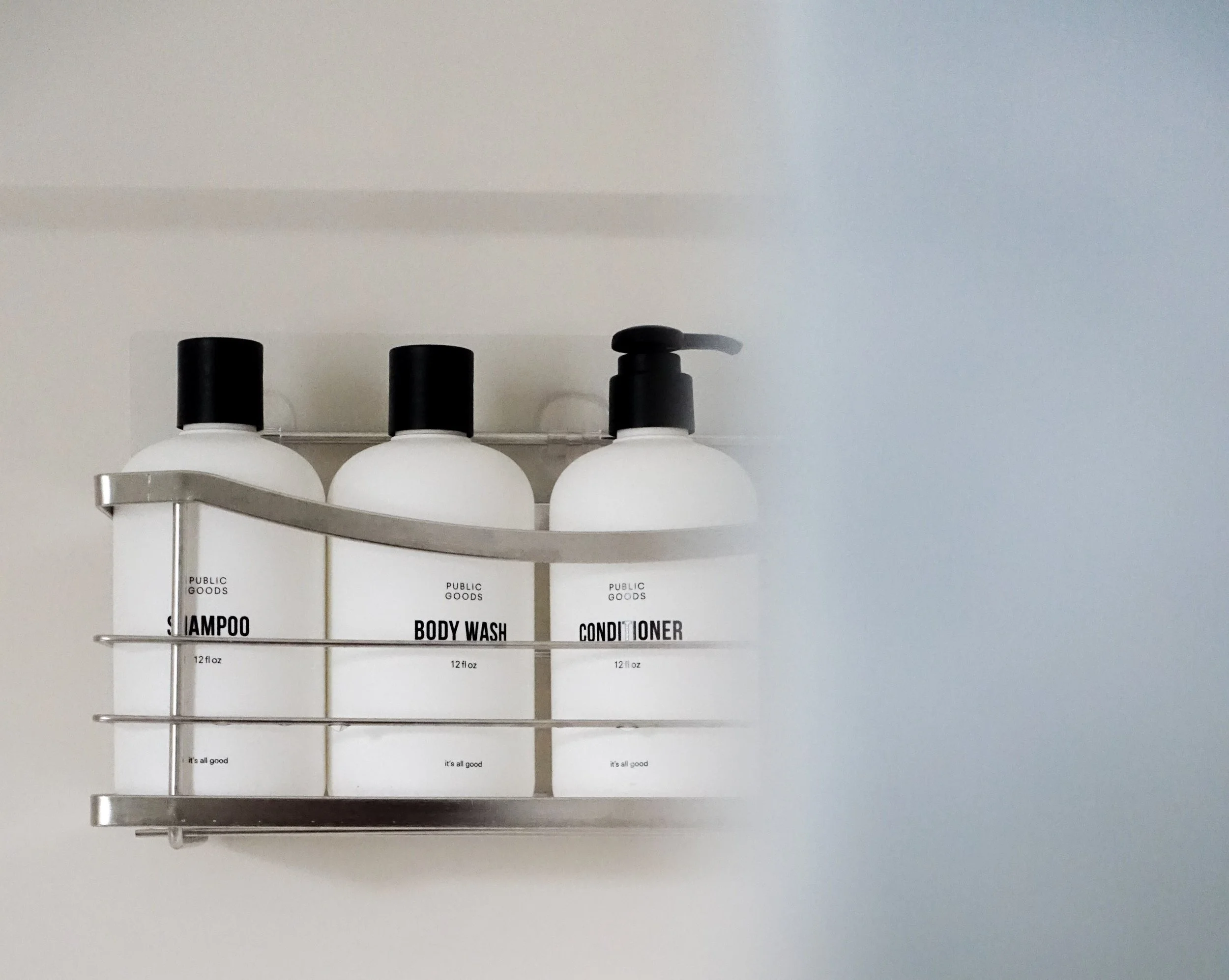 Three bottles of public goods shampoo, body wash, and conditioner on a wall-mounted metal shelf, with a blurred background.