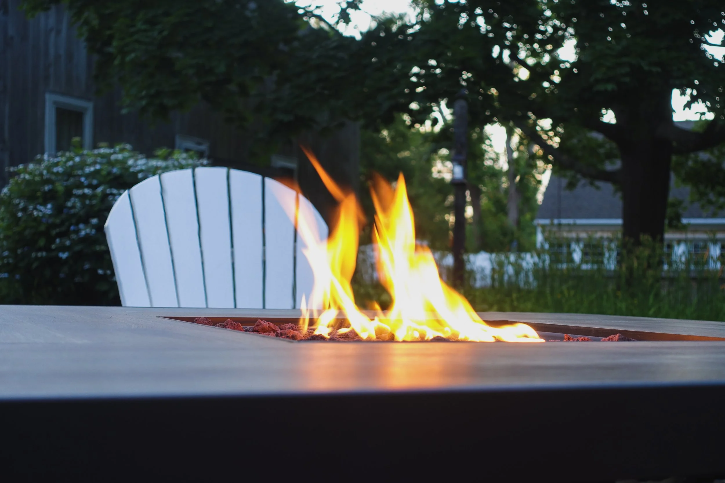 Outdoor fire pit with flames on a patio table in a backyard with trees and a house in the background.