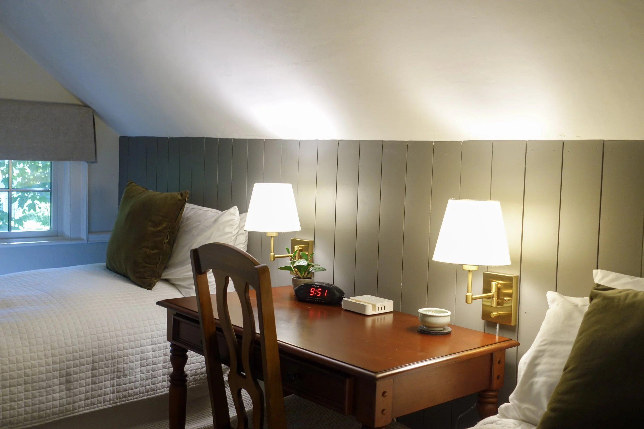 A cozy hotel room with two beds, a wooden nightstand between them, two white lamps, a digital alarm clock, and a window with blinds.