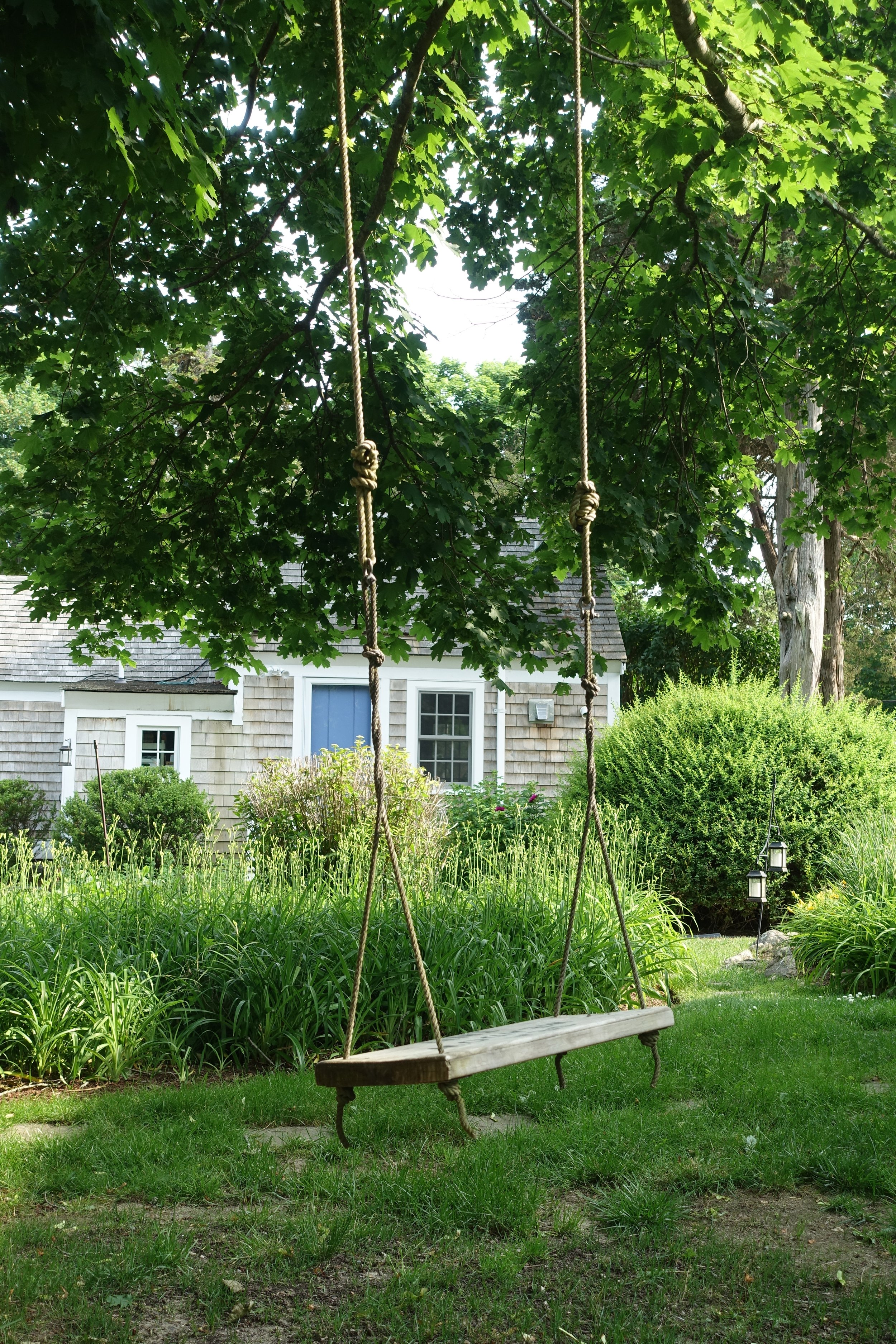 A wooden swing hanging from a tree in a garden, with a house and lush greenery in the background.