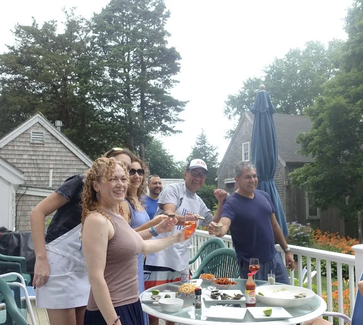 Group of six people enjoying a backyard gathering, standing around a grill and table with food, outdoors with trees, houses, and a large blue umbrella.