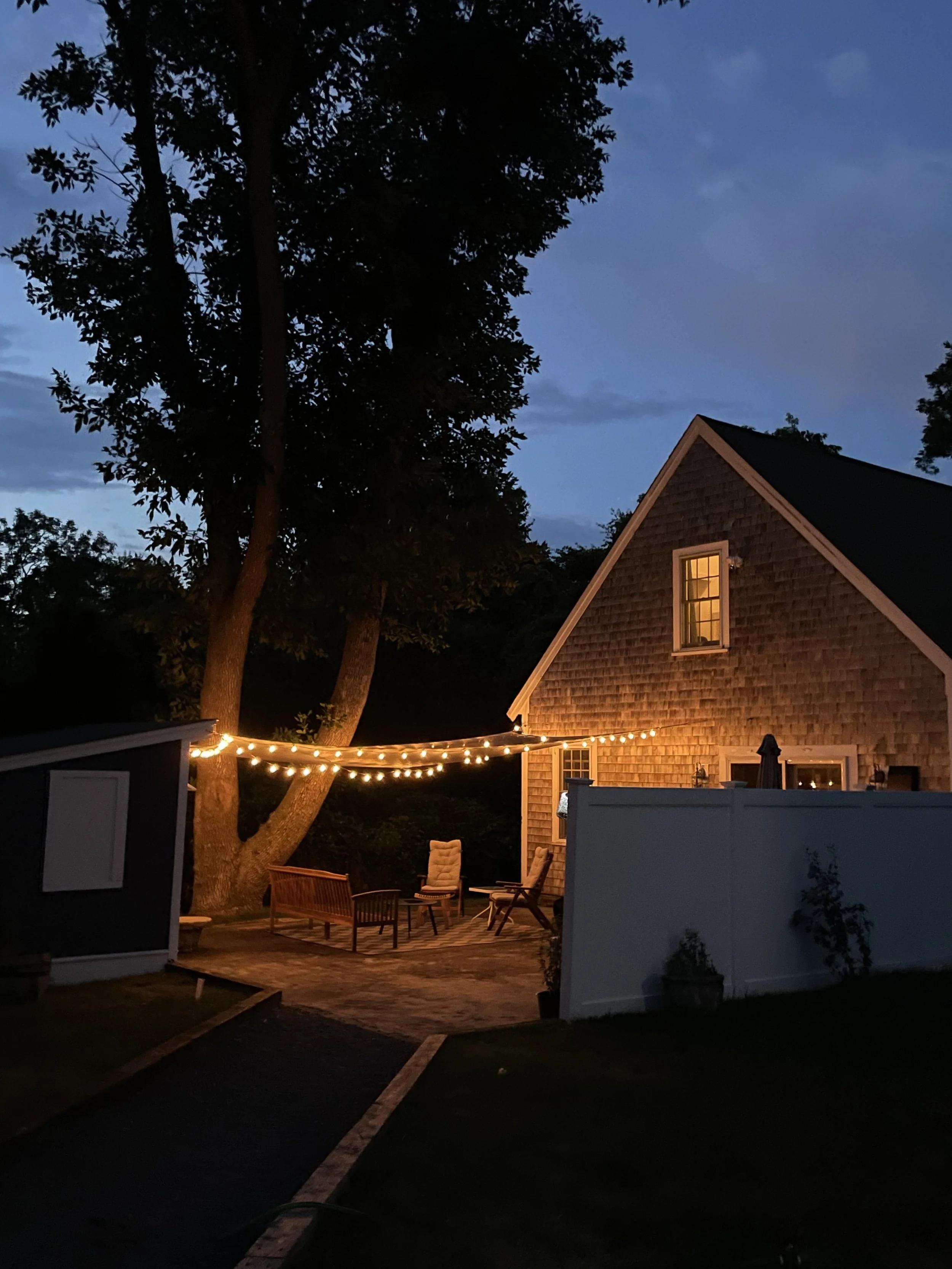 An outdoor patio with string lights, chairs, a bench, and a house with windows lit from inside. Tall trees with partly cloudy sky in the background during evening.