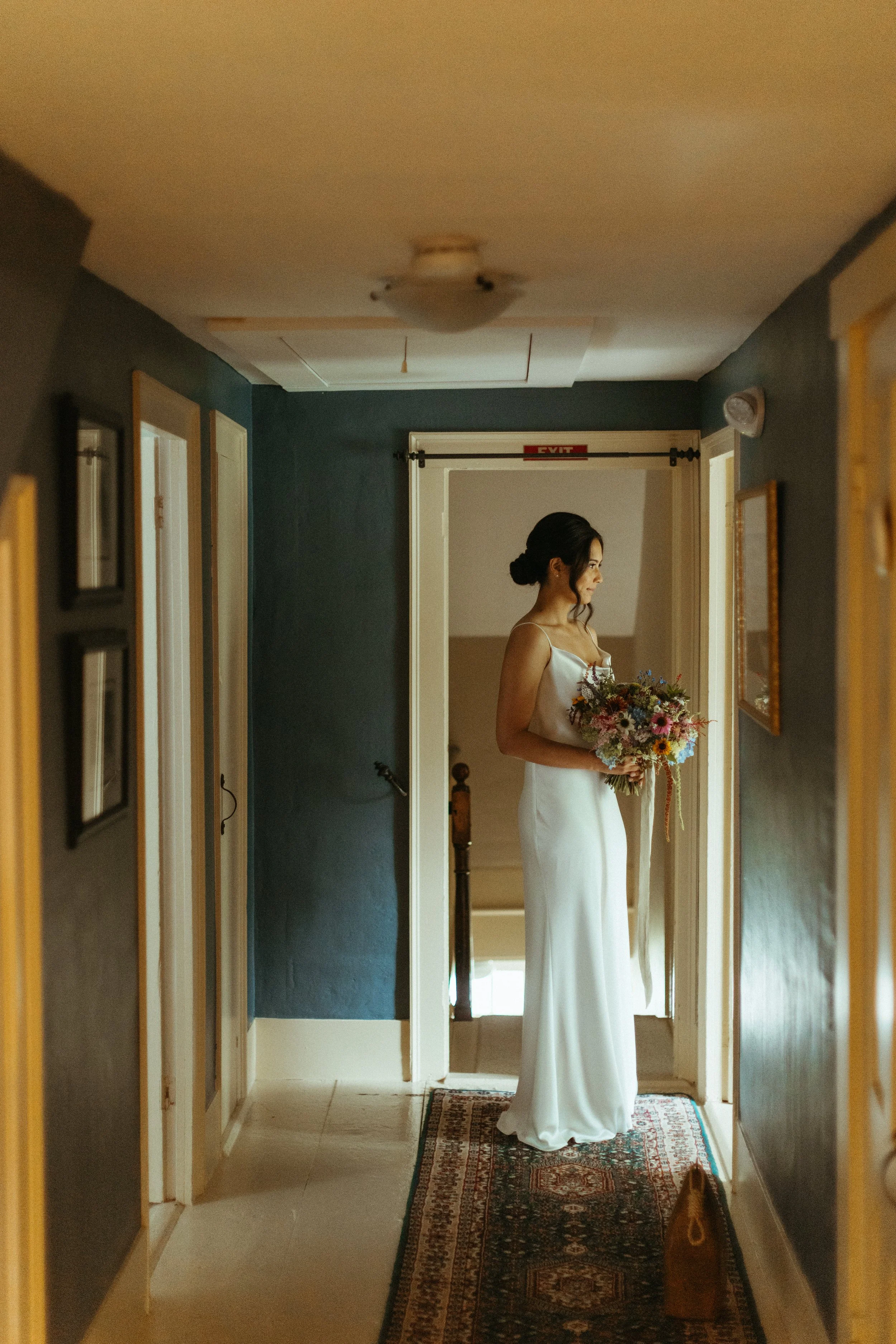 A woman in a white dress holding a bouquet, standing in a hallway next to a doorway with an 'exit' sign above it.