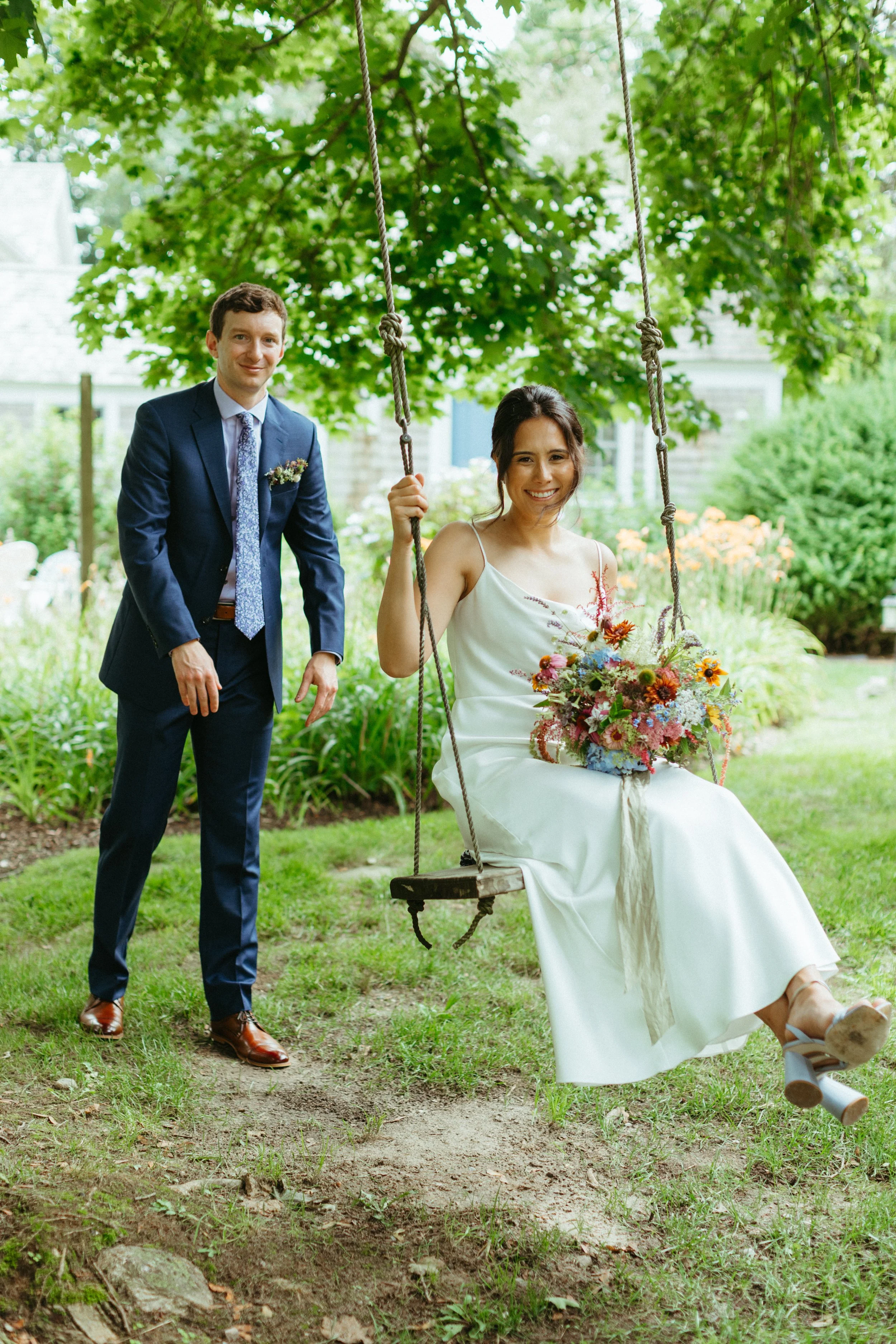 A woman in a white dress sitting on a swing holding a colorful bouquet of flowers, with a man in a blue suit standing nearby, in a garden setting.