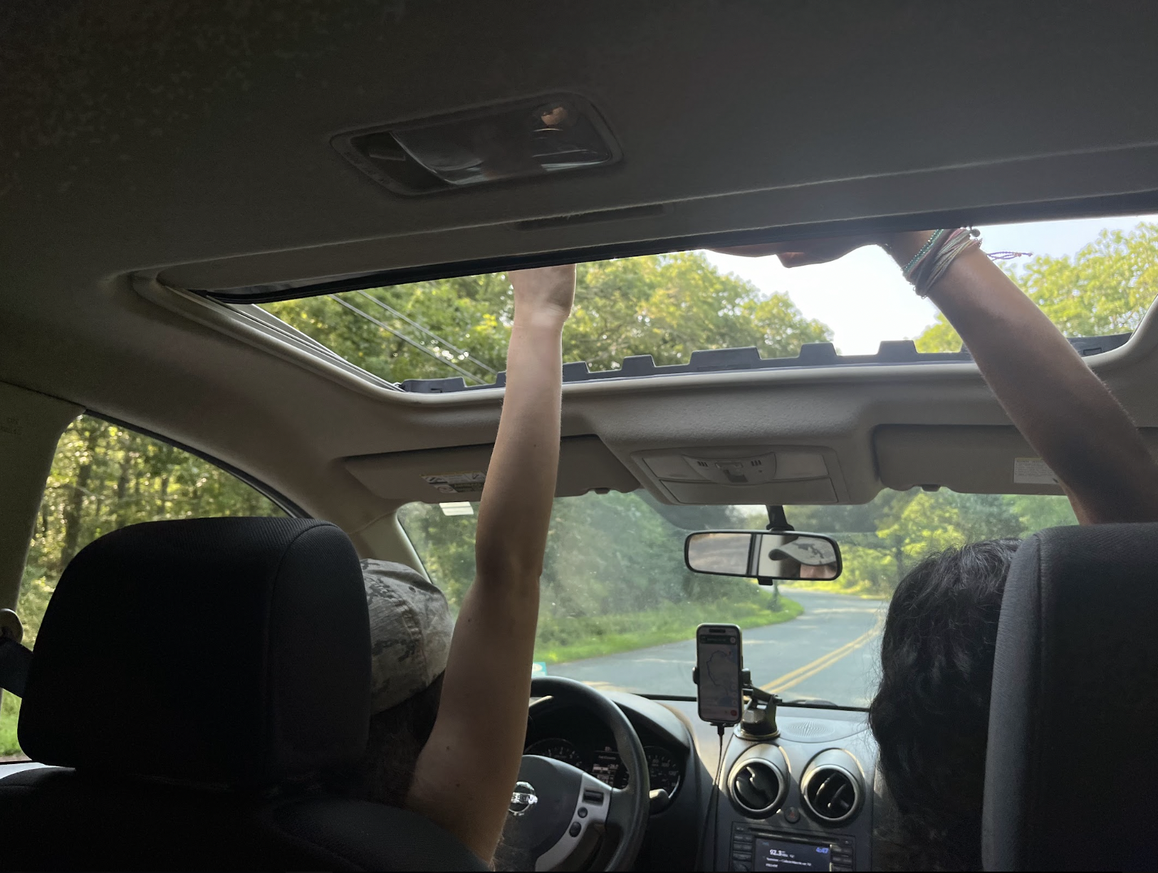 Inside a car, with two people in the front seats. The driver is holding their hand up and touching the open sunroof, while the passenger is looking out the window. The car is on a road with green trees visible outside.