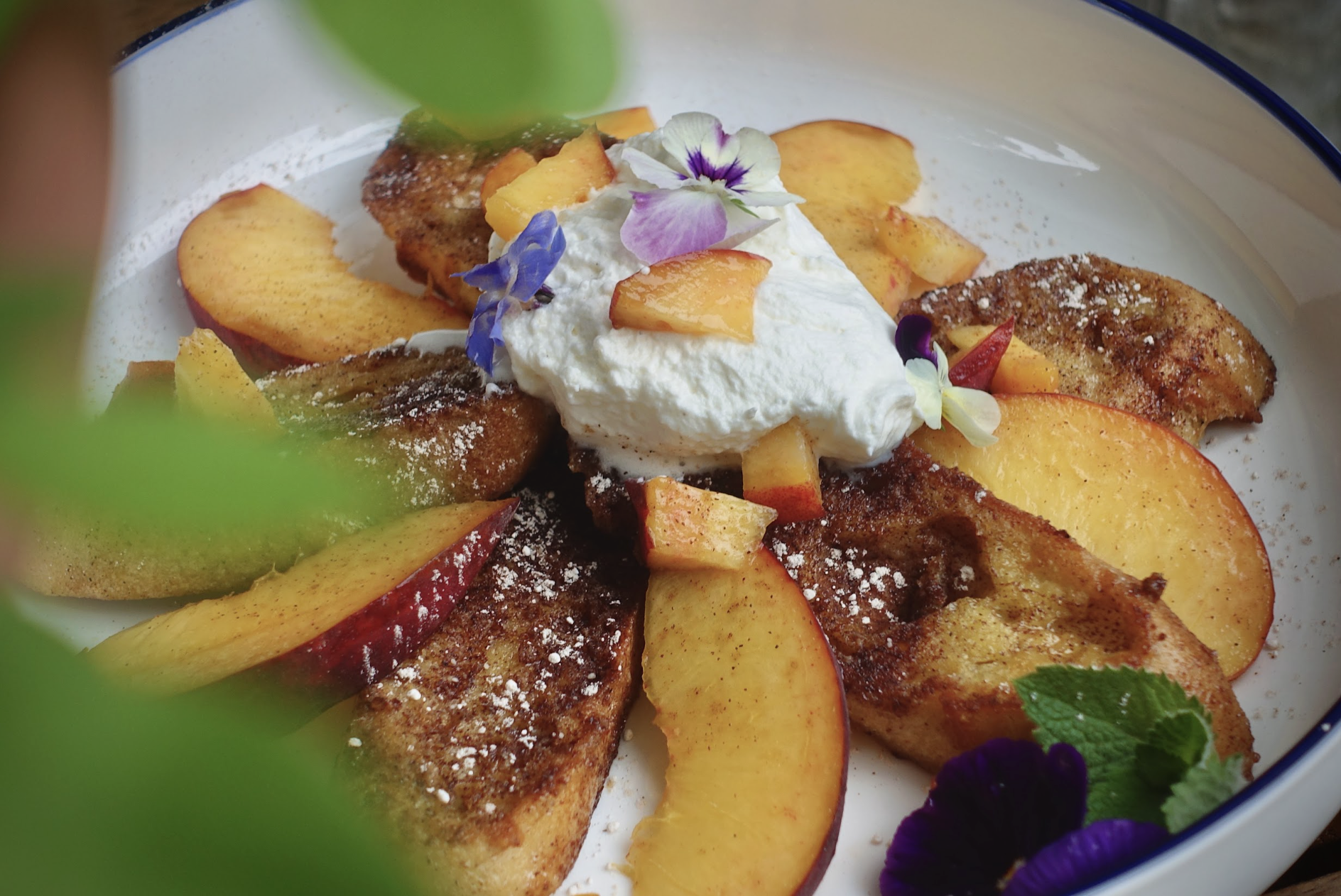 Sliced peaches and french toast garnished with edible flowers, whipped cream, and powdered sugar, served on a white plate with a blue rim.