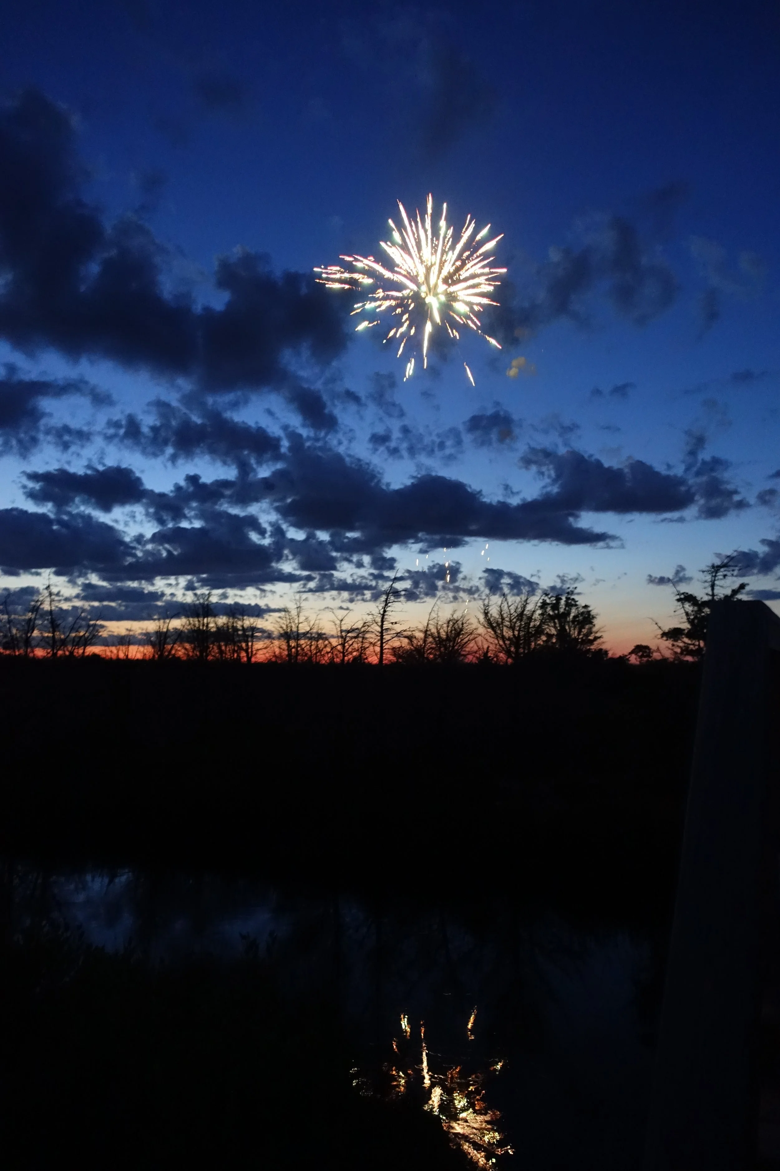 Fireworks explode in the evening sky, with dark clouds and a sunset in the background, reflected in a body of water below.