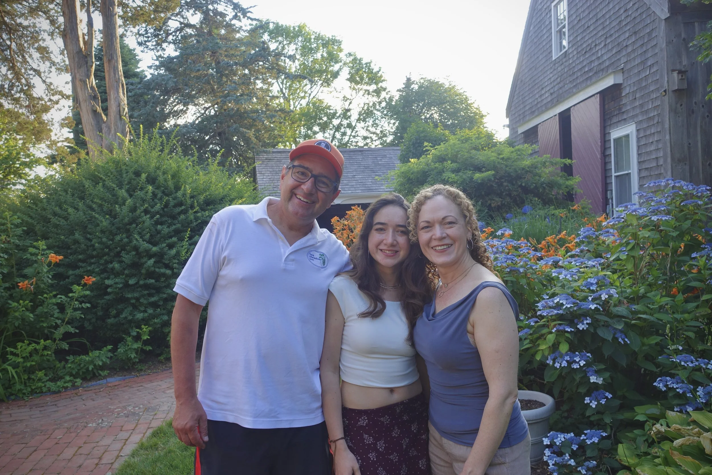 Three people smiling and standing together in a garden with trees, bushes, and blue flowers, during daytime.