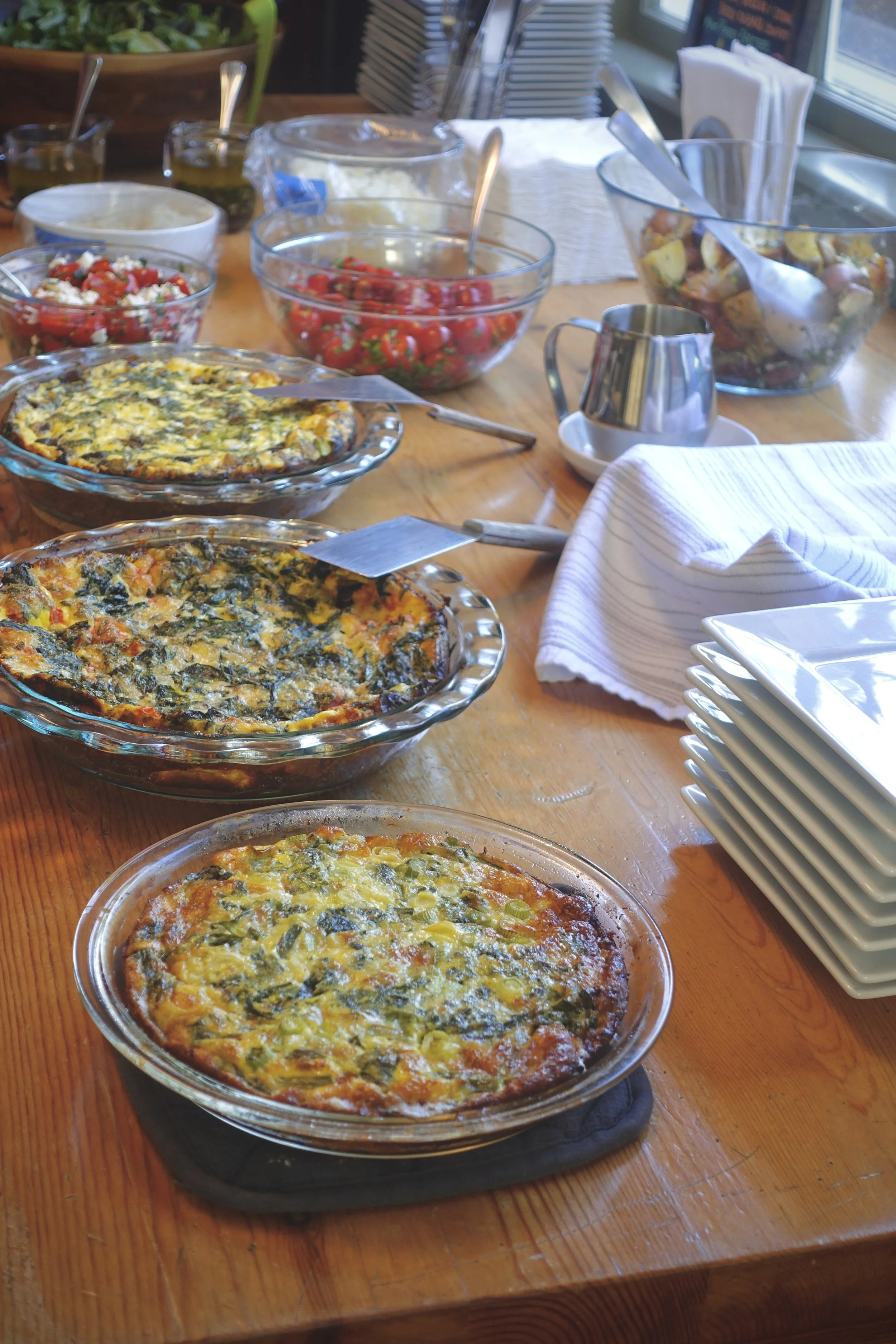 Various vegetable and spinach quiches on a wooden table, with salads and bowls in the background.