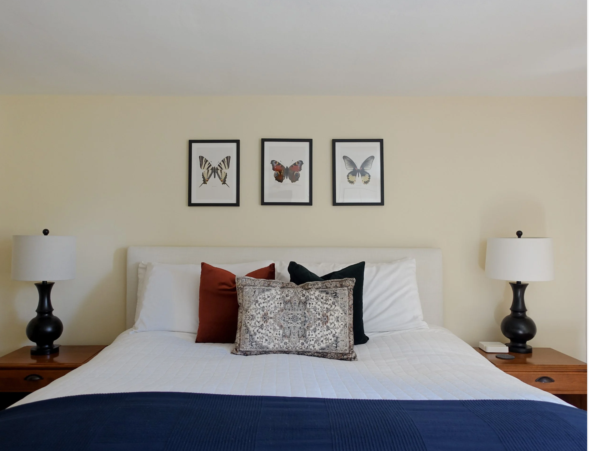 A neatly made bed with a white bedspread, accented by four throw pillows in maroon, patterned brown, and black, is flanked by two black bedside lamps on wooden nightstands. Above the bed are three framed butterfly illustrations on a beige wall.