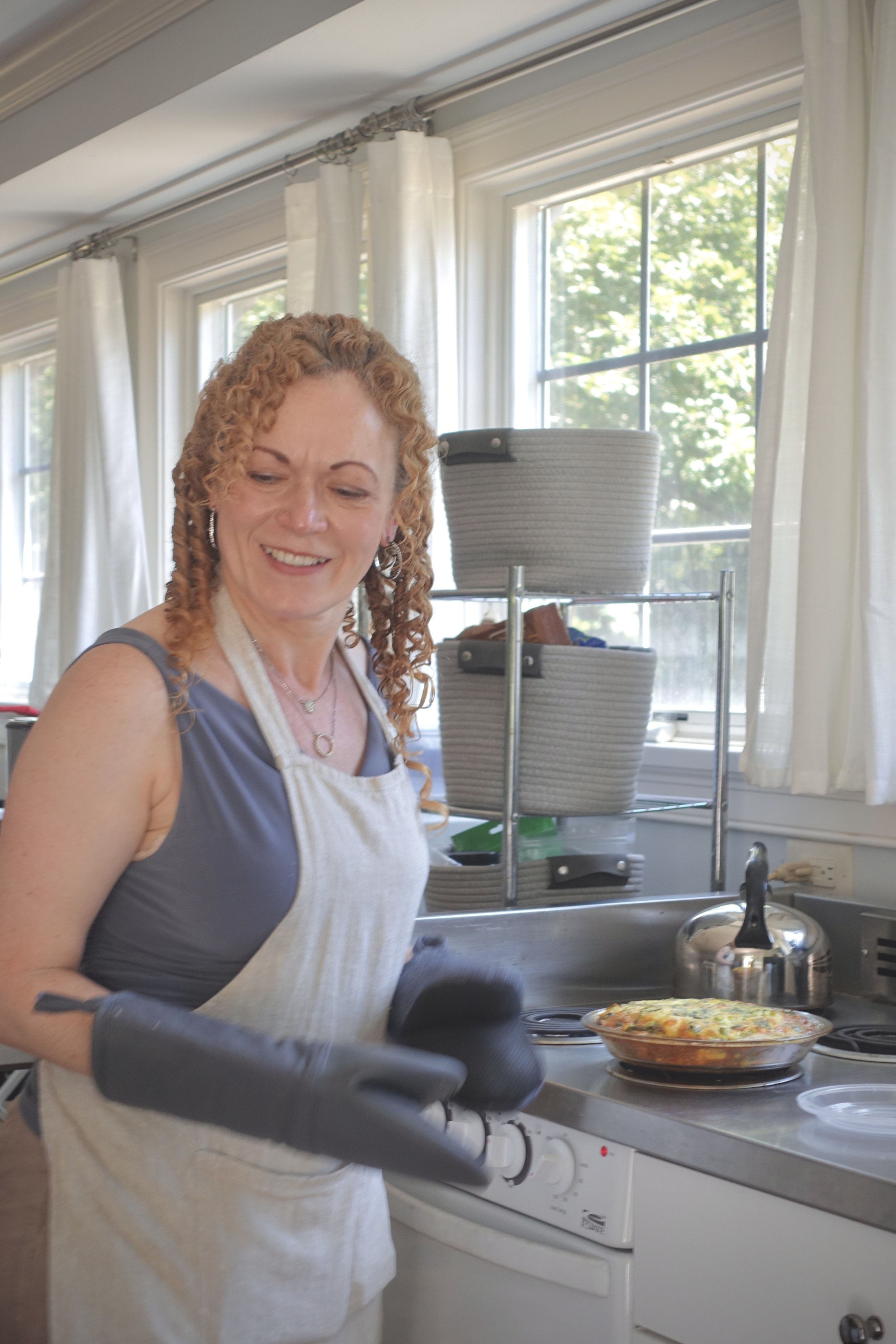 A woman with curly red hair wearing a gray shirt and beige apron cooking a dish in the kitchen, with white curtains and windows showing trees outside.