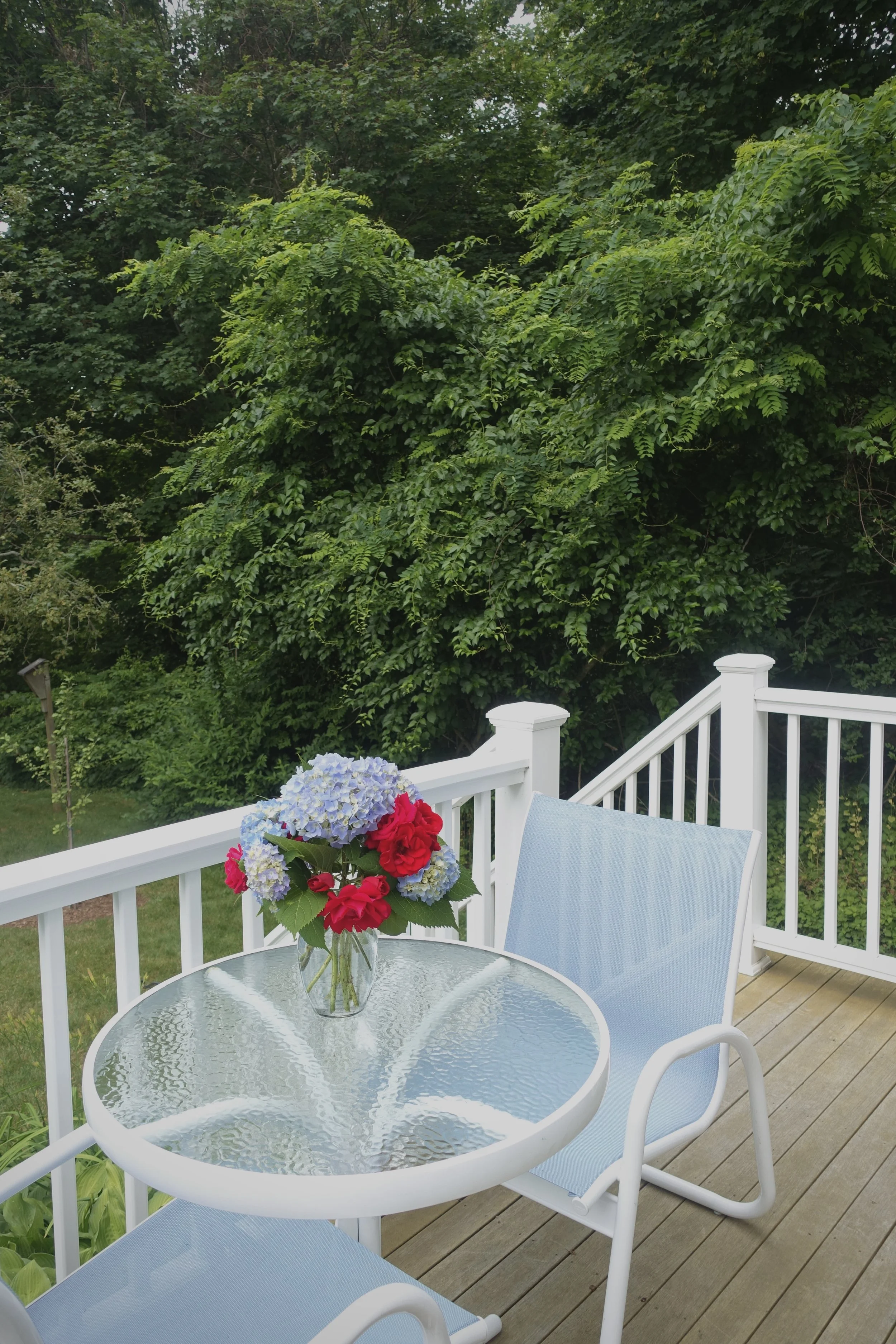 View of an outdoor deck with a glass-top table and two light blue chairs, decorated with a vase of hydrangeas and red flowers, surrounded by green trees and a white railing.