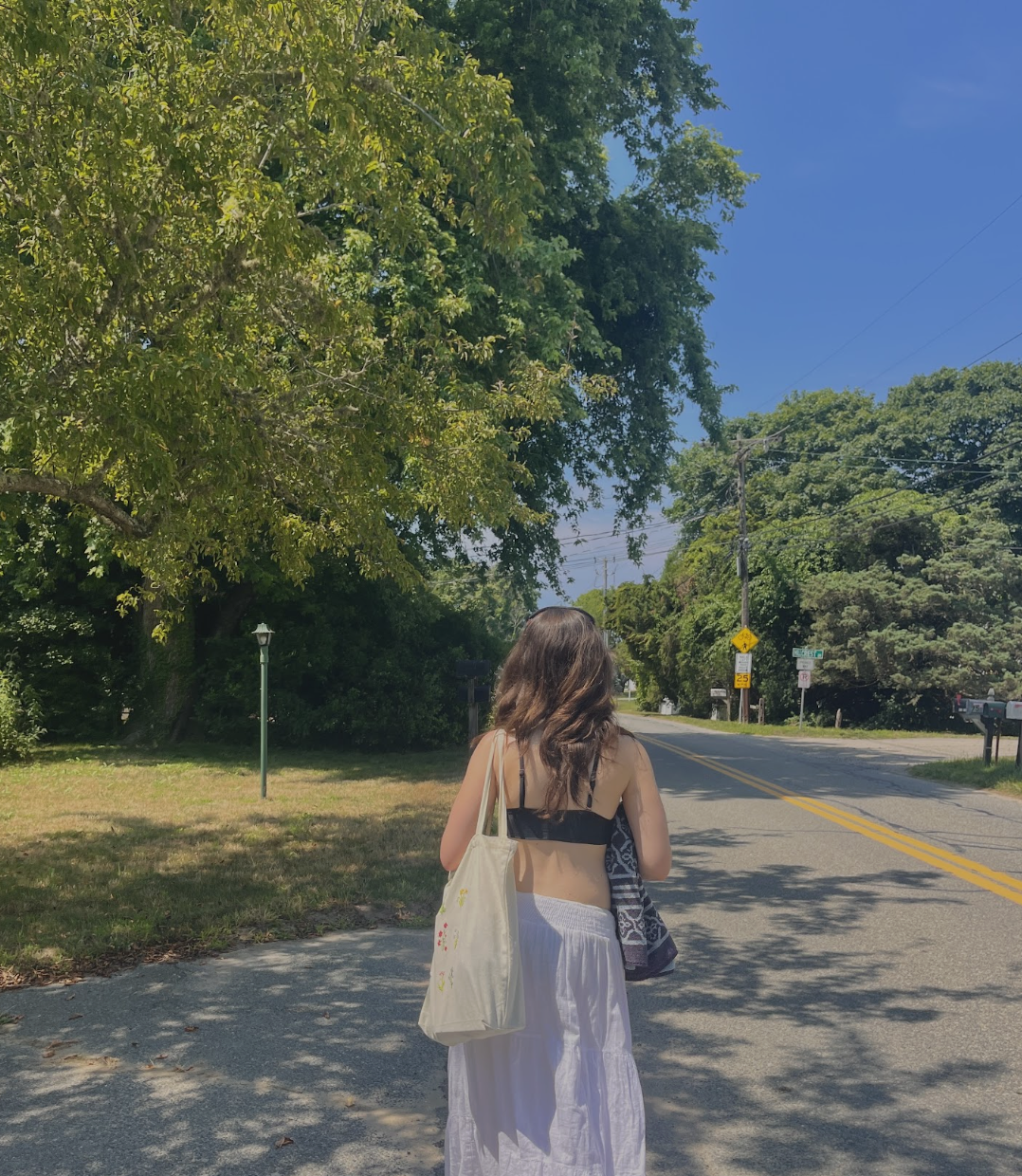 A woman with long brown hair, wearing a black crop top and white maxi skirt, is walking on the sidewalk with a beige tote bag and a towel or blanket in her hand. She is walking away from the camera along a suburban street lined with trees and utility