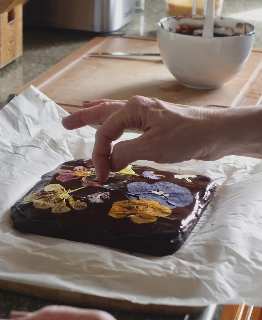 A person is decorating a piece of chocolate with dried flower petals on parchment paper, with bowls and utensils on a wooden table nearby.