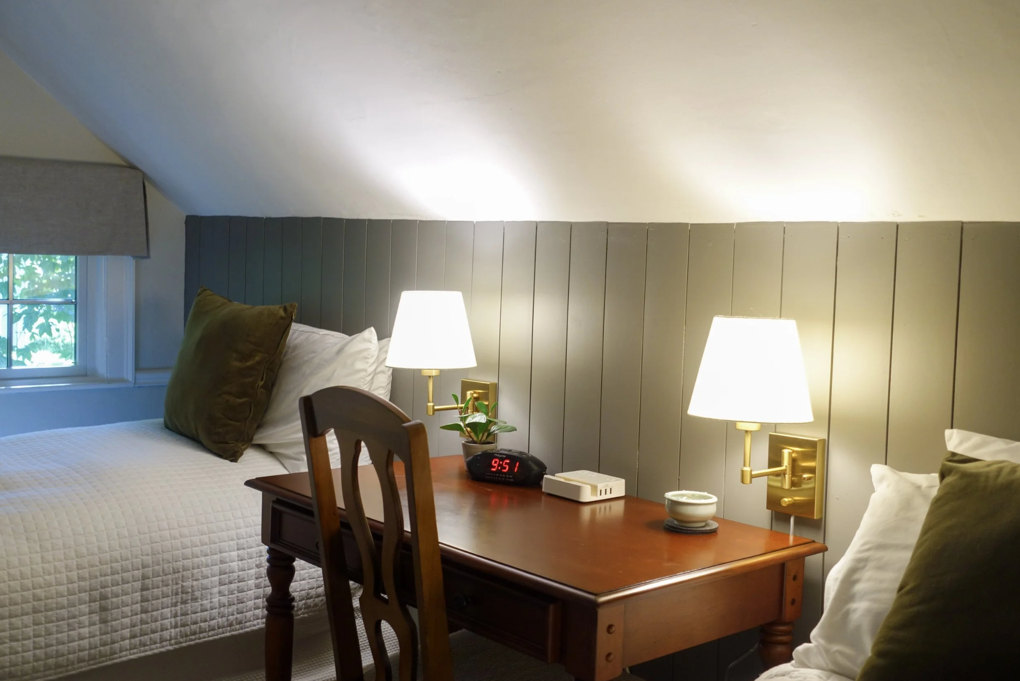 A bedroom with a bed, two pillows, a bedside table with a lamp, an alarm clock showing 9:51, a small plant, a tissue box, and a window with a green view. The wall behind the bed has vertical paneling, and there are two matching lamps on either side.