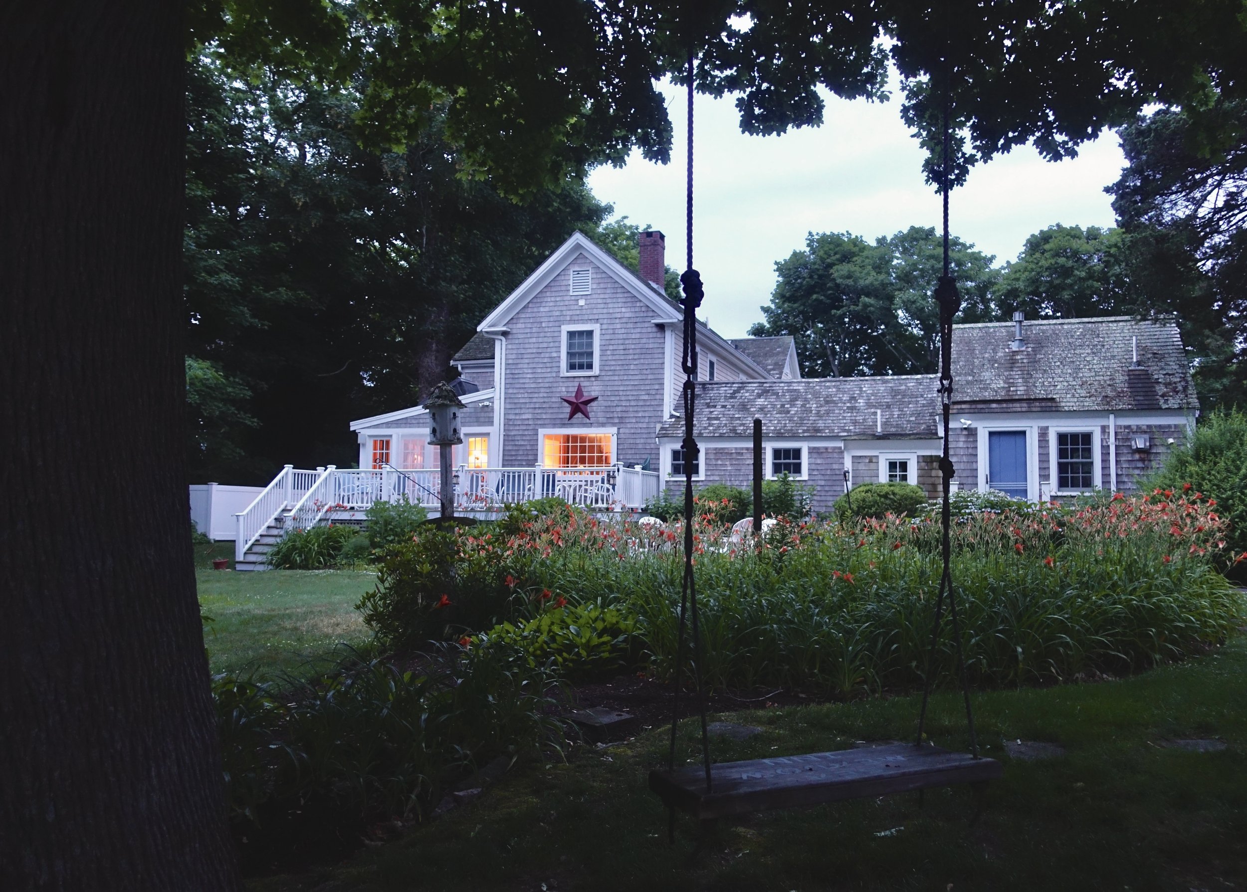 A house with gray wooden siding and a steep roof, viewed from a garden with a swing in the foreground, trees surrounding the house, warm interior lights visible through the windows, a small porch with a white railing, and a star decoration on the hou