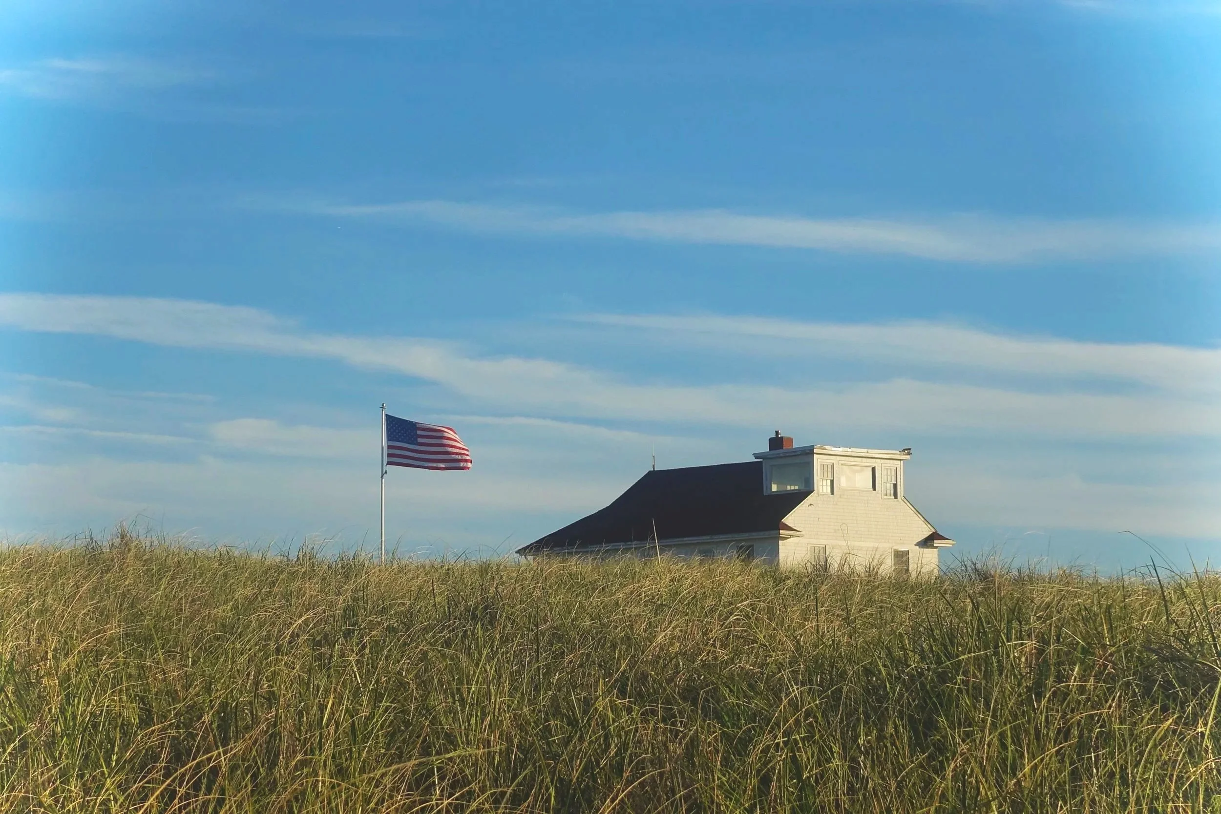 A white house with a red roof in a grassy field under a blue sky with wispy clouds, and an American flag flying on a pole nearby.