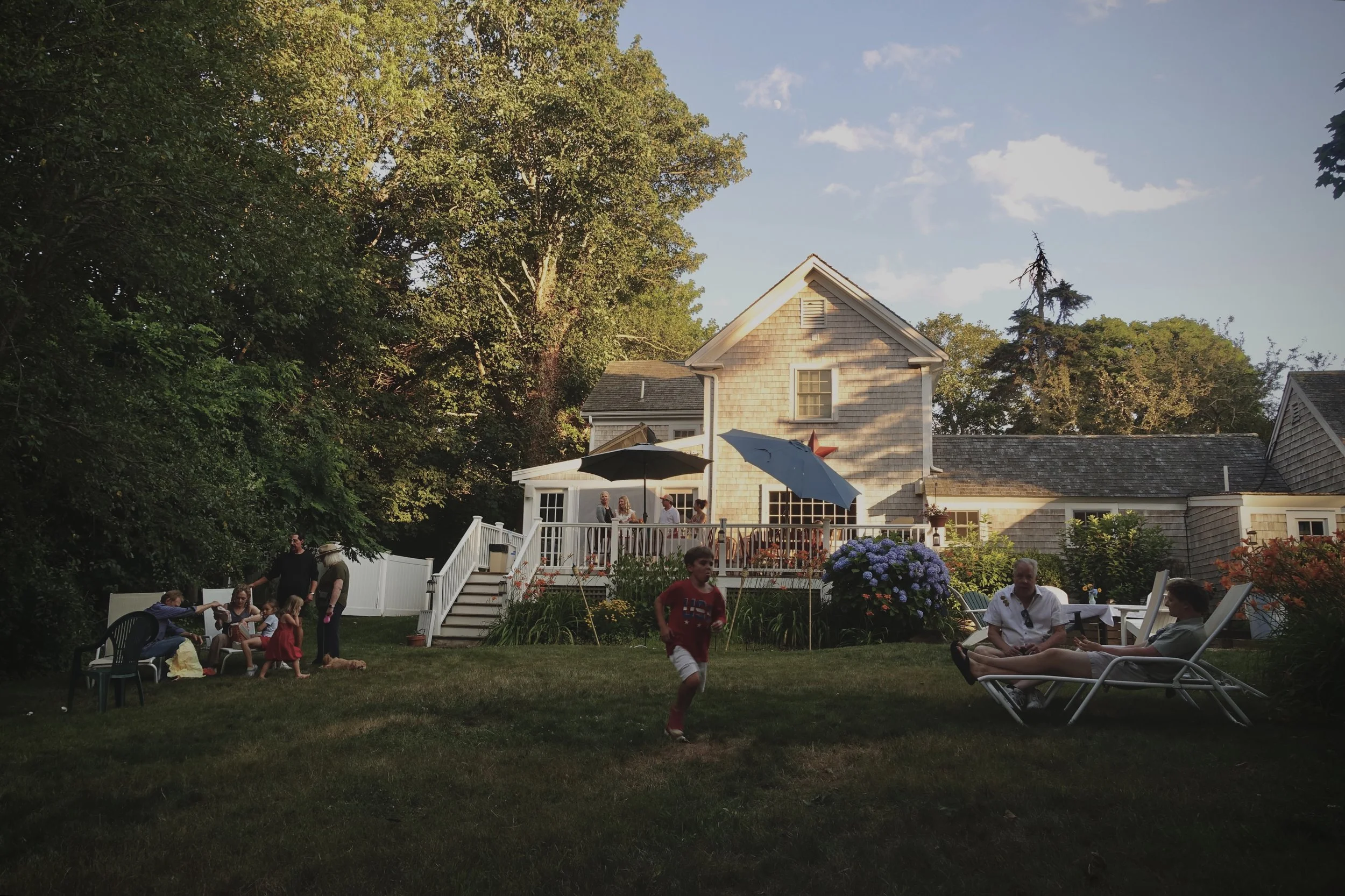 A backyard gathering with people enjoying a sunny evening, children playing on the lawn, and adults sitting and chatting near a house with a deck decorated with umbrellas and flowers.