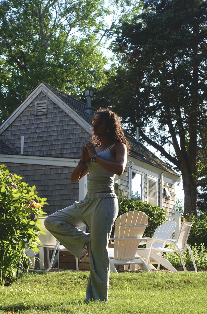 A woman practicing yoga outdoors in a garden, with a house and trees in the background.