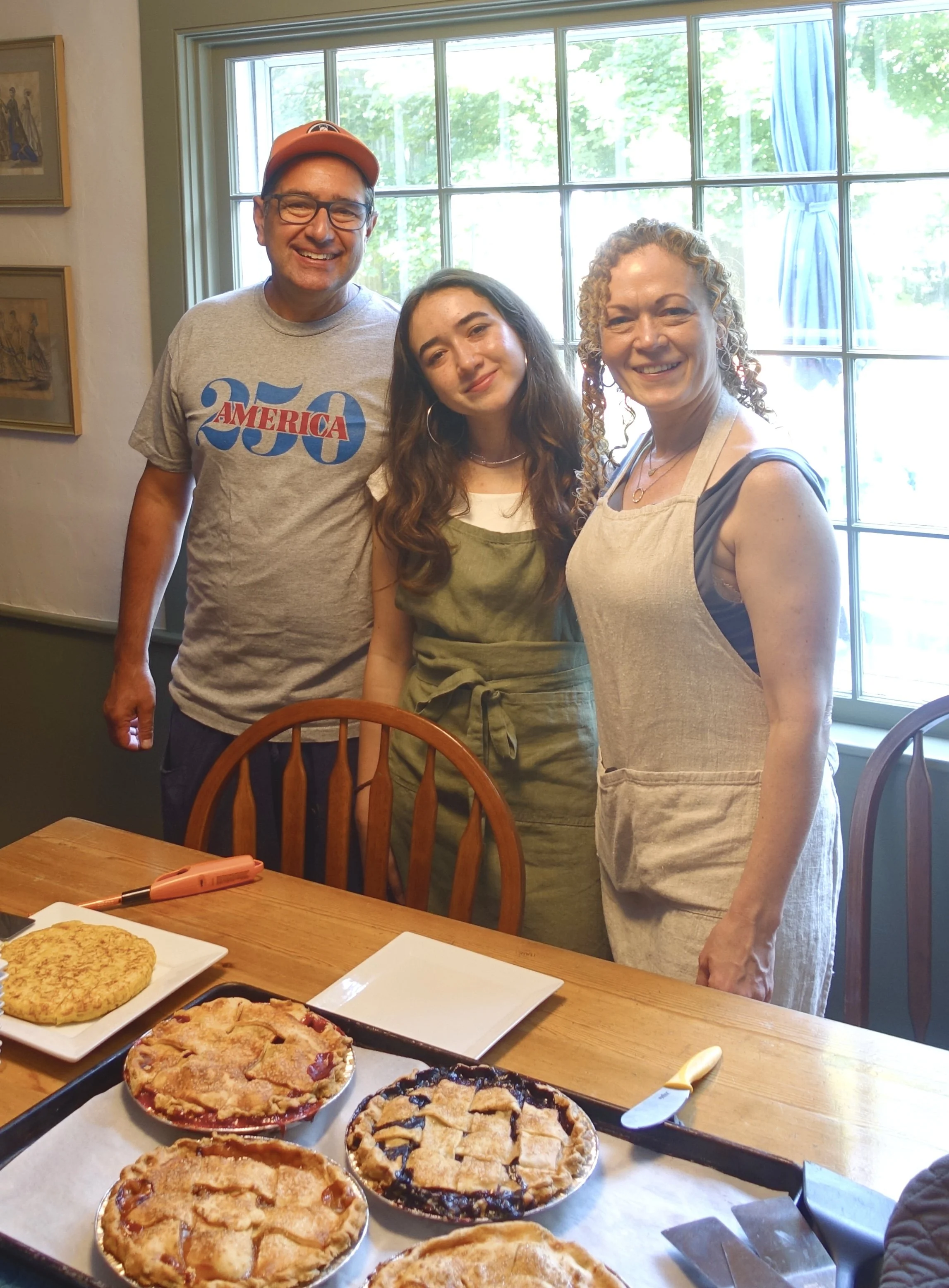 Three people standing around a table with baked pies, smiling inside a house with a large window and green trees outside.