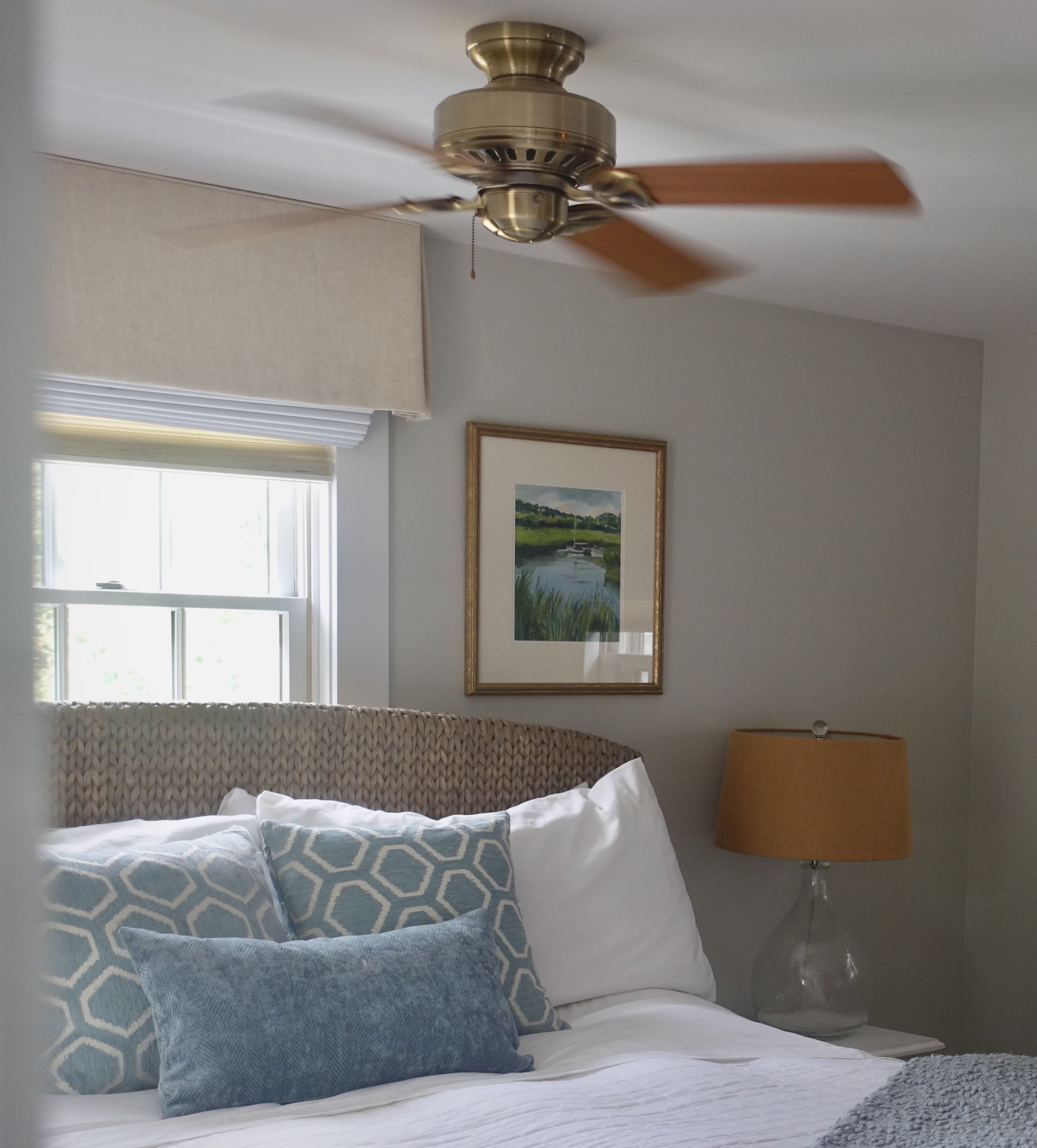 A bedroom with a bed that has a woven headboard, two gray pillows with geometric patterns, a light-colored blanket, and a gray throw at the foot. A window with blinds and a framed landscape painting are on the wall, and there is a nightstand with a lamp beside the bed.