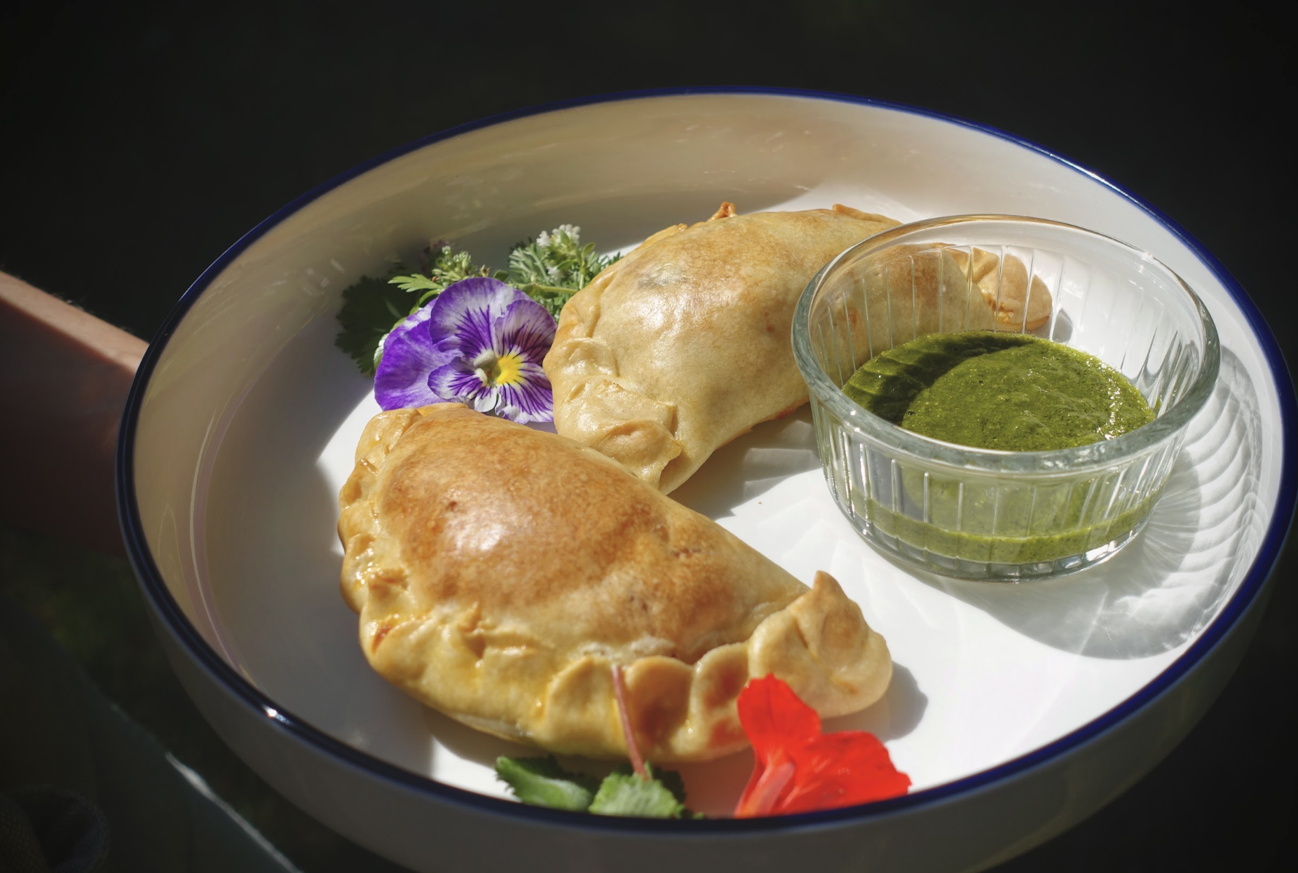 A white plate with a blue rim containing two golden-brown empanadas, a small glass bowl of green dipping sauce, a purple edible flower, and some green herbs, with a small red flower at the bottom.