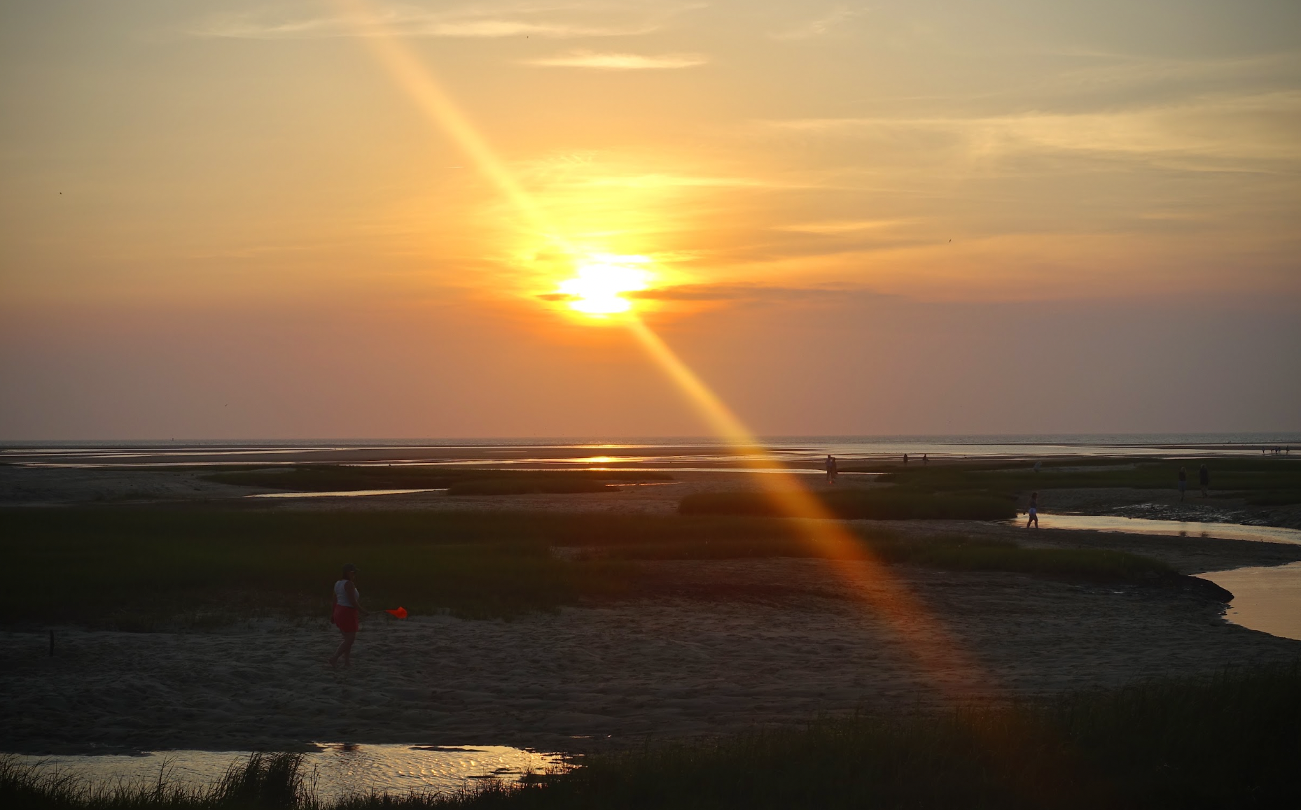 Sunset over a beach with a bright sun and a rainbow-like rainbow, people walking on the sand and near the water, grassy areas and water channels.