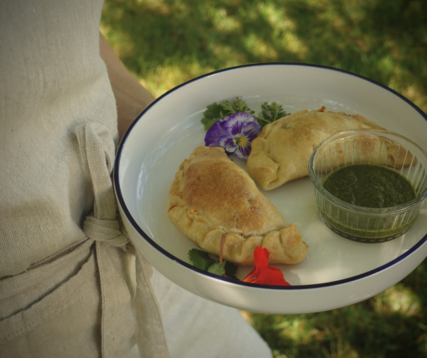Person holding a white plate with two empanadas, purple and yellow flower, green cilantro garnish, and a small glass bowl of green sauce, outdoors with green grass background.