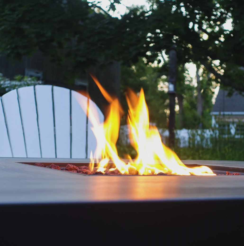 Outdoor table with a fire feature, flames burning among rocks, surrounded by chairs and trees.