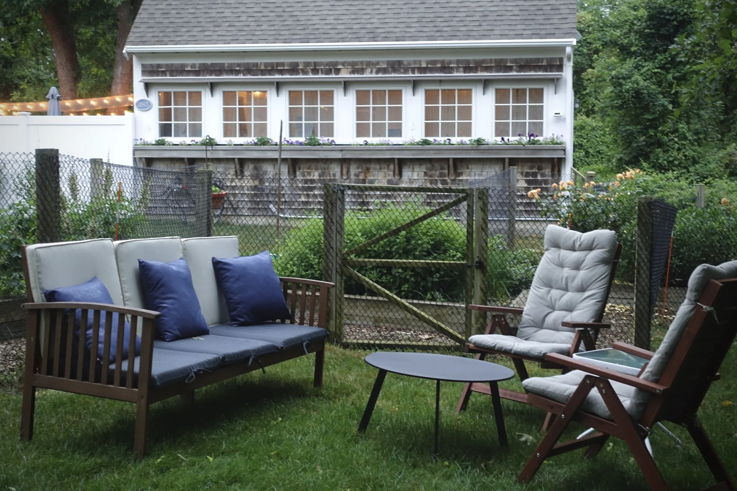 A backyard patio with outdoor seating, including a cushioned wooden bench with blue pillows, a white cushioned chair, and a wooden chair, with a small round table. In the background, there is a fenced garden, a shed with windowed doors, and lush gree