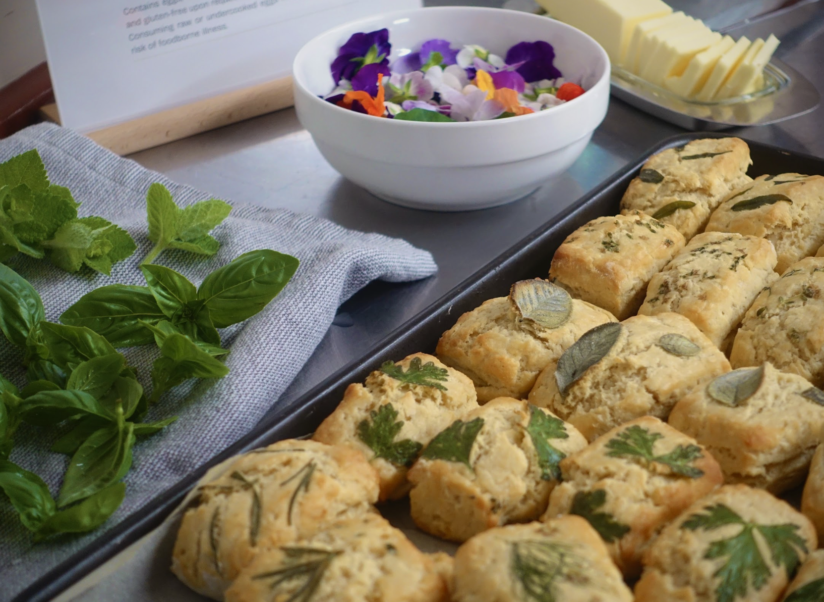 Freshly baked herb biscuits topped with sage and parsley, placed on a baking tray. Surrounding garnishes include basil and mint leaves, with a bowl of edible flowers, slices of cheese, and a sign in the background.