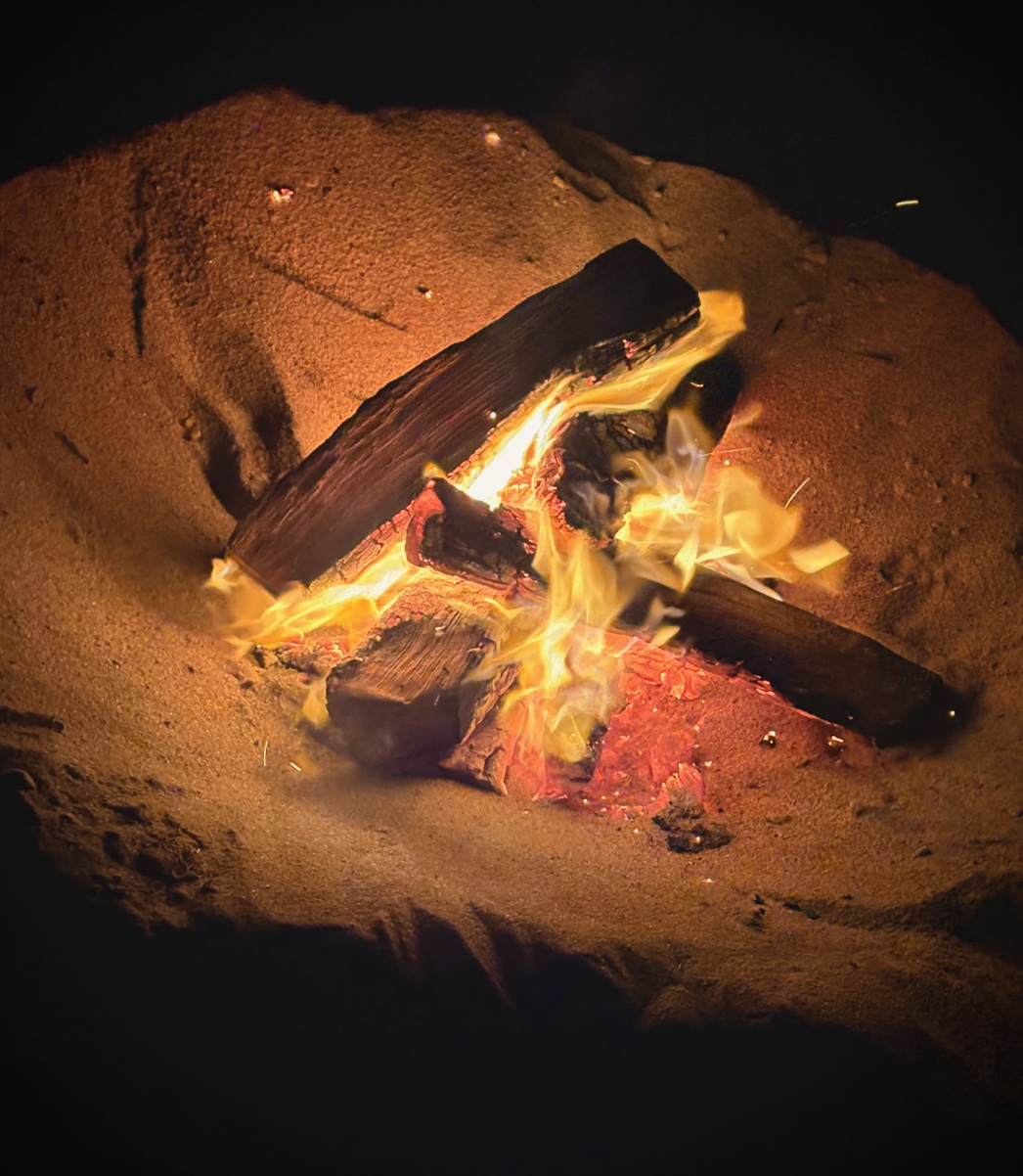 A small campfire burning in a fire pit or sand, with glowing embers and flames around the logs.