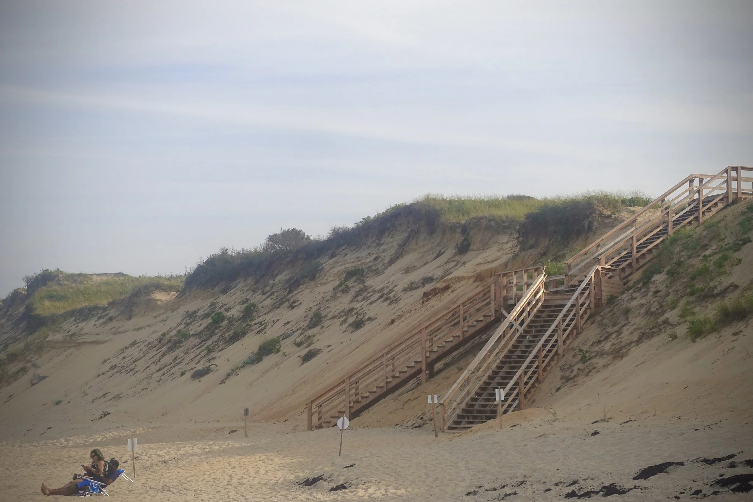Beach scene with wooden stairs leading up a sandy cliff, a woman sitting on the sand reading a book, and Japanese signs on the beach.