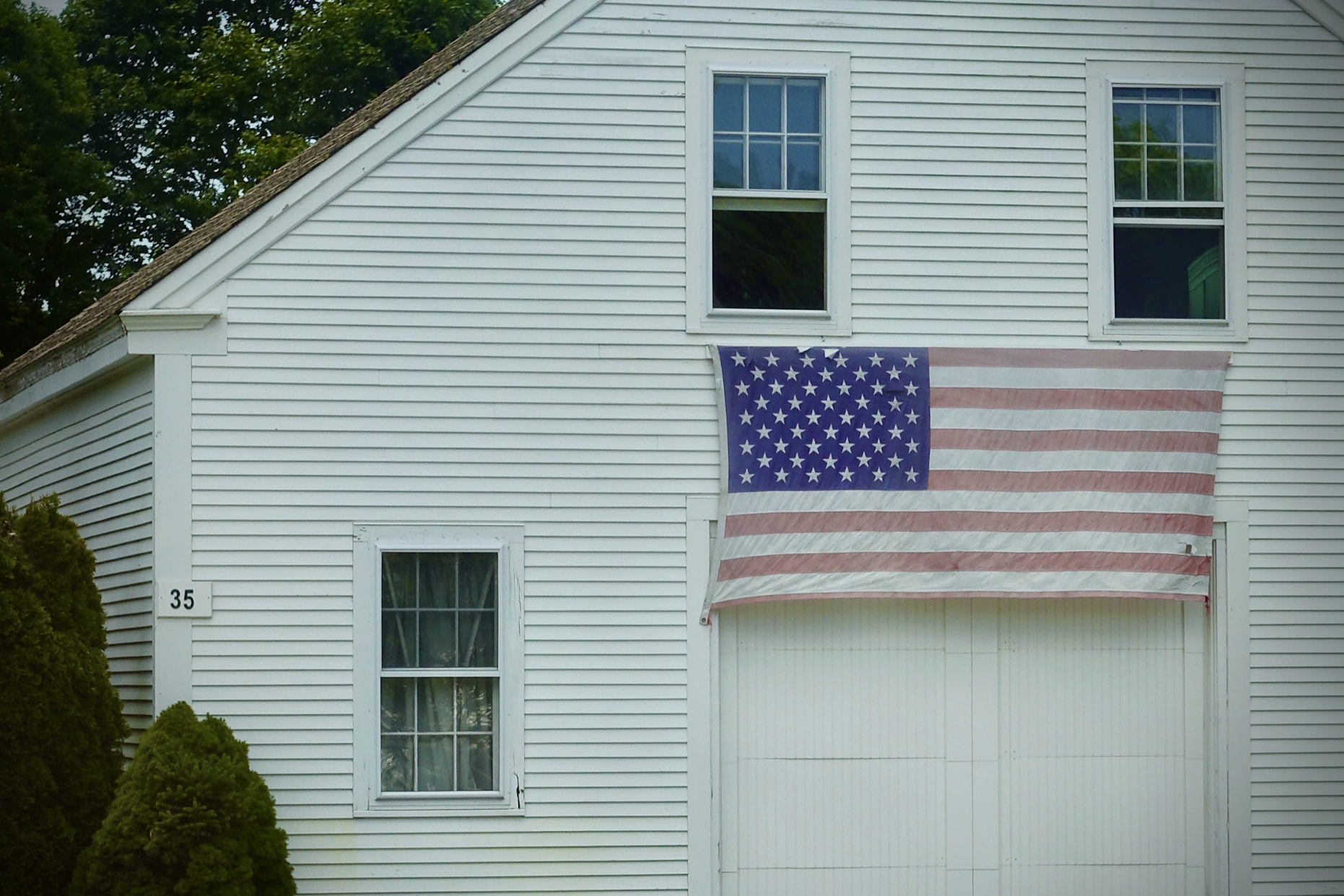 White house with vertical siding, three windows, and an American flag hanging on the front. The house number 35 is visible next to the lower left window. Green trees and bushes are around the house.