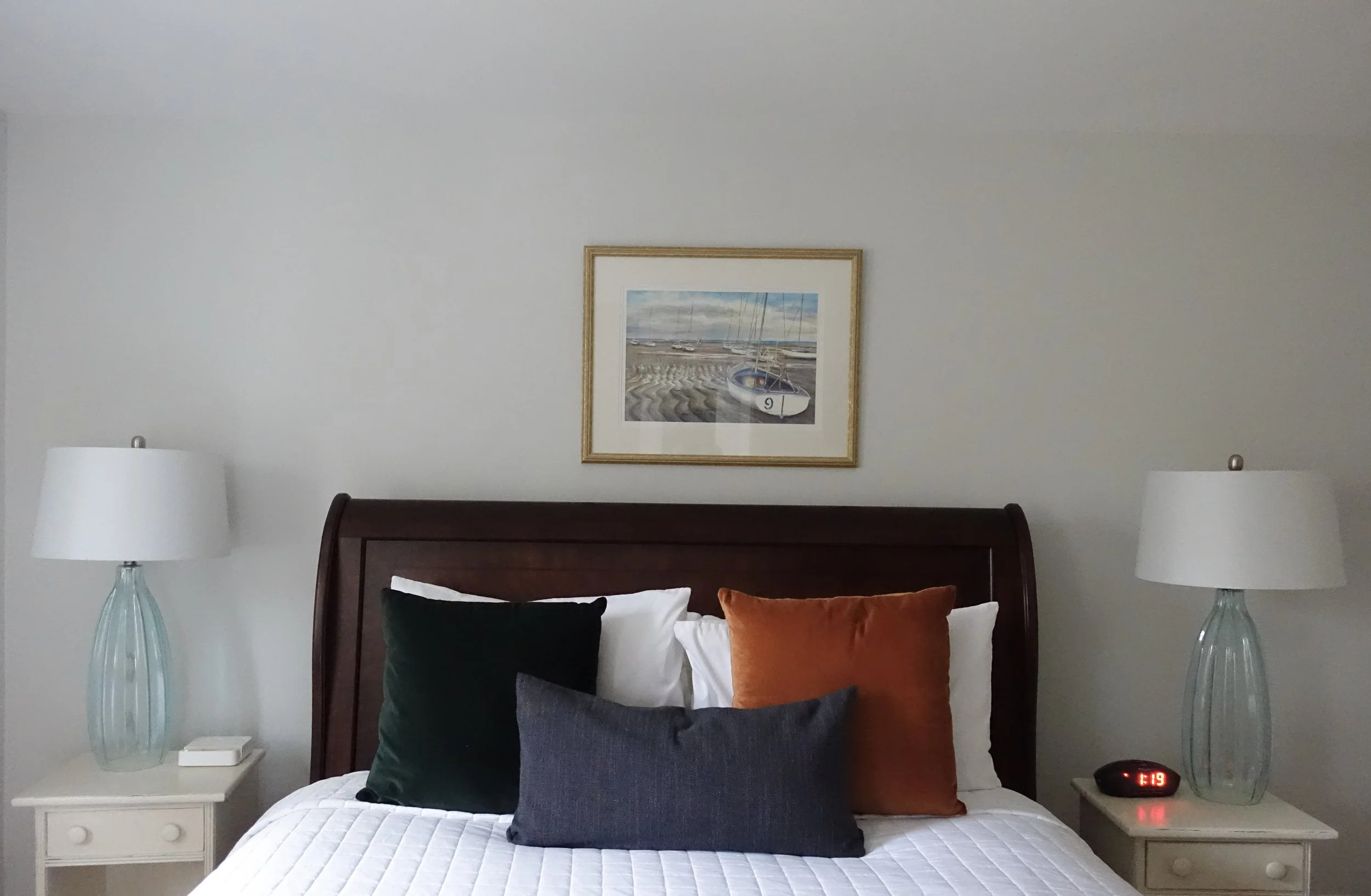 A neatly made bed in a bedroom with a wooden headboard, colorful pillows, and two matching lamps on white nightstands. A framed seaside painting hangs above.