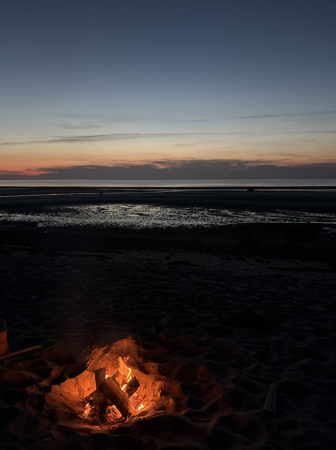 A beach at sunset with a small campfire burning in the sand in the foreground, and the ocean and colorful sky in the background.