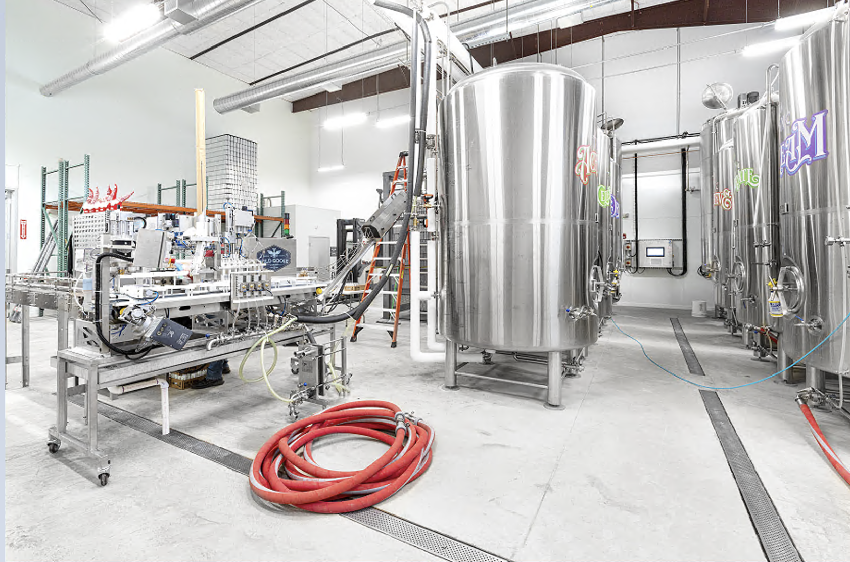 Inside a brewery or water processing facility with large stainless steel tanks, pipes, and equipment for brewing or filtration, and a coiled red hose on the floor.