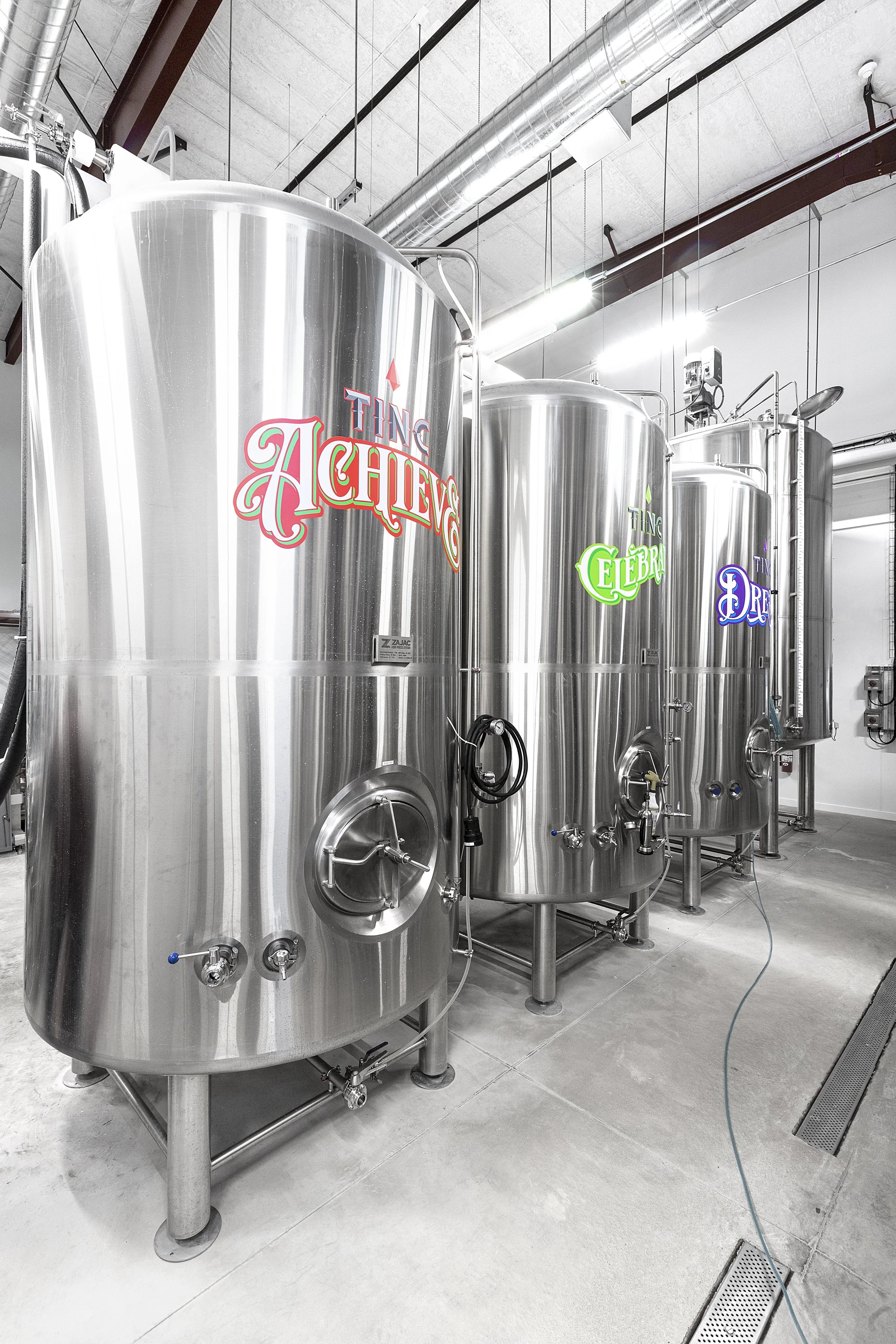 Industrial brewery tanks made of stainless steel in a brewery or manufacturing facility, with labels reading 'Ting Aching,' 'Ting Celebrate,' and 'Ting Dream'.