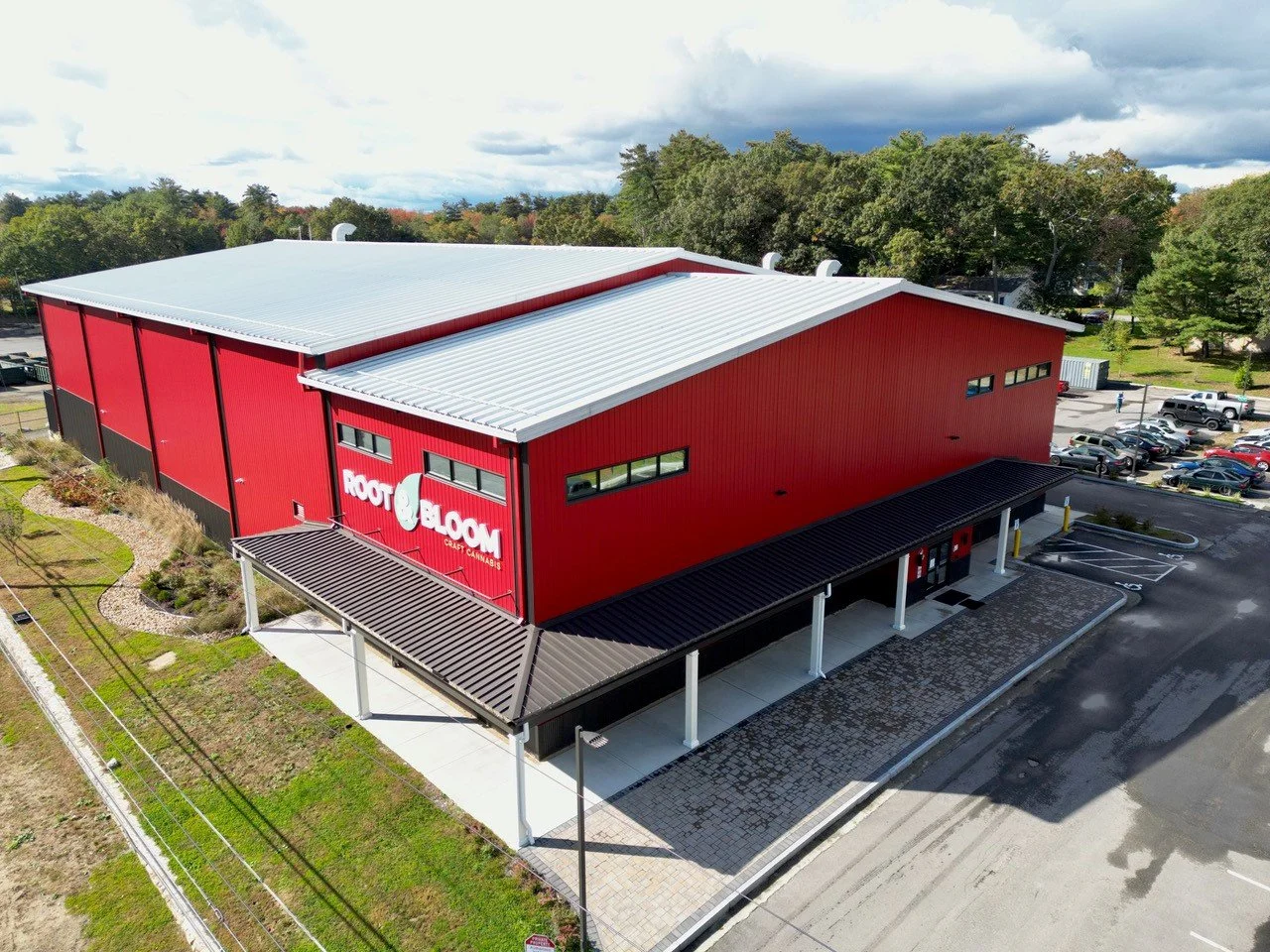 An aerial view of a red building labeled 'Rooted Bloom Craft Cannabis' with a parking lot and green trees in the background.