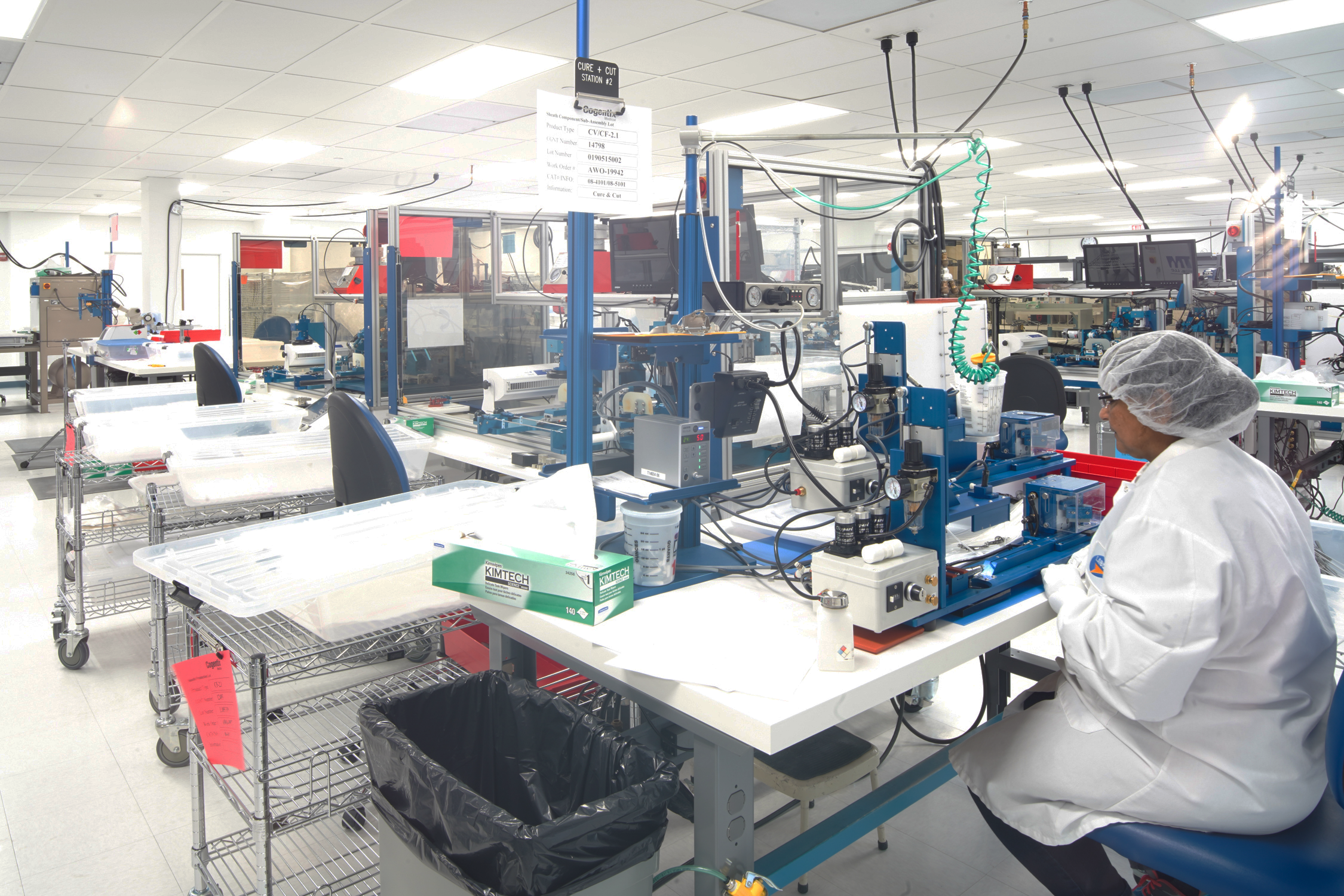 A worker in a cleanroom setting wearing a white lab coat, hairnet, and glasses, working with medical or laboratory equipment on a workbench with various tools, monitors, and carts around.