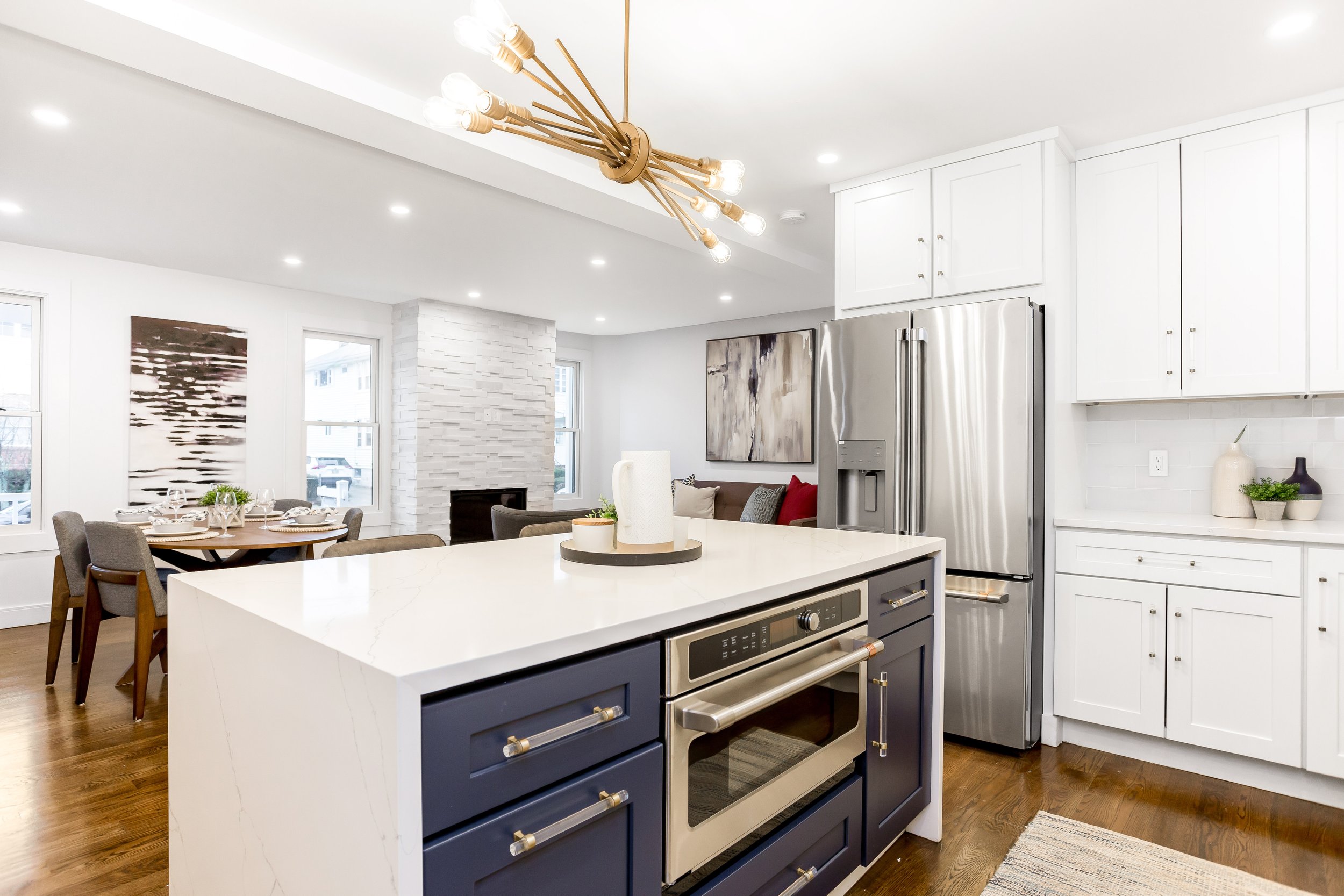 Modern kitchen with white cabinets, stainless steel refrigerator, and a large kitchen island with a white countertop and navy blue drawers. There is a dining area with a wooden table and gray chairs, a white brick fireplace, and abstract artwork on the walls. A gold and glass chandelier hangs from the ceiling, and hardwood floors complete the space.