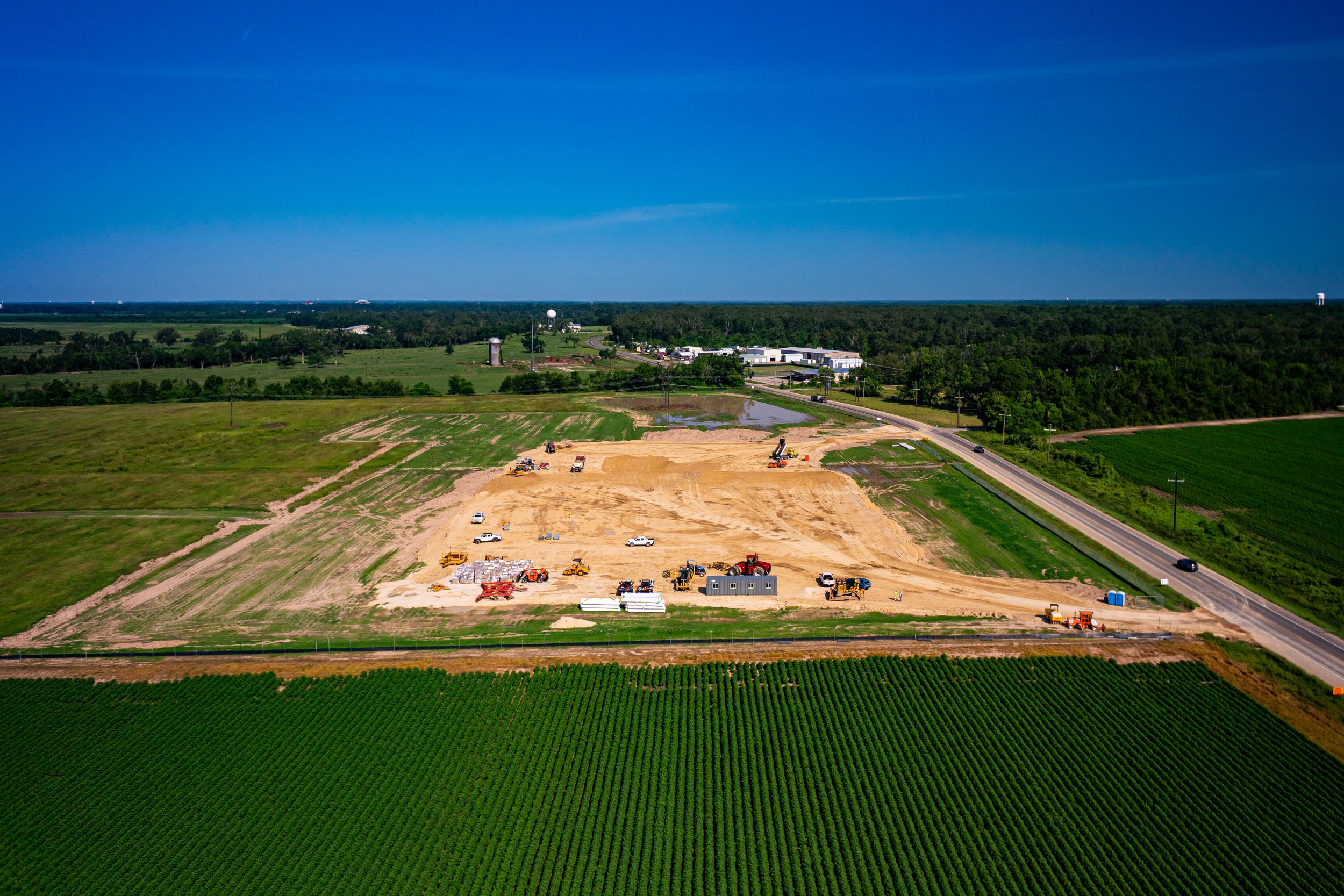 An aerial view of farmland and construction site with equipment and vehicles, green fields in the foreground, and a blue sky with a few clouds.