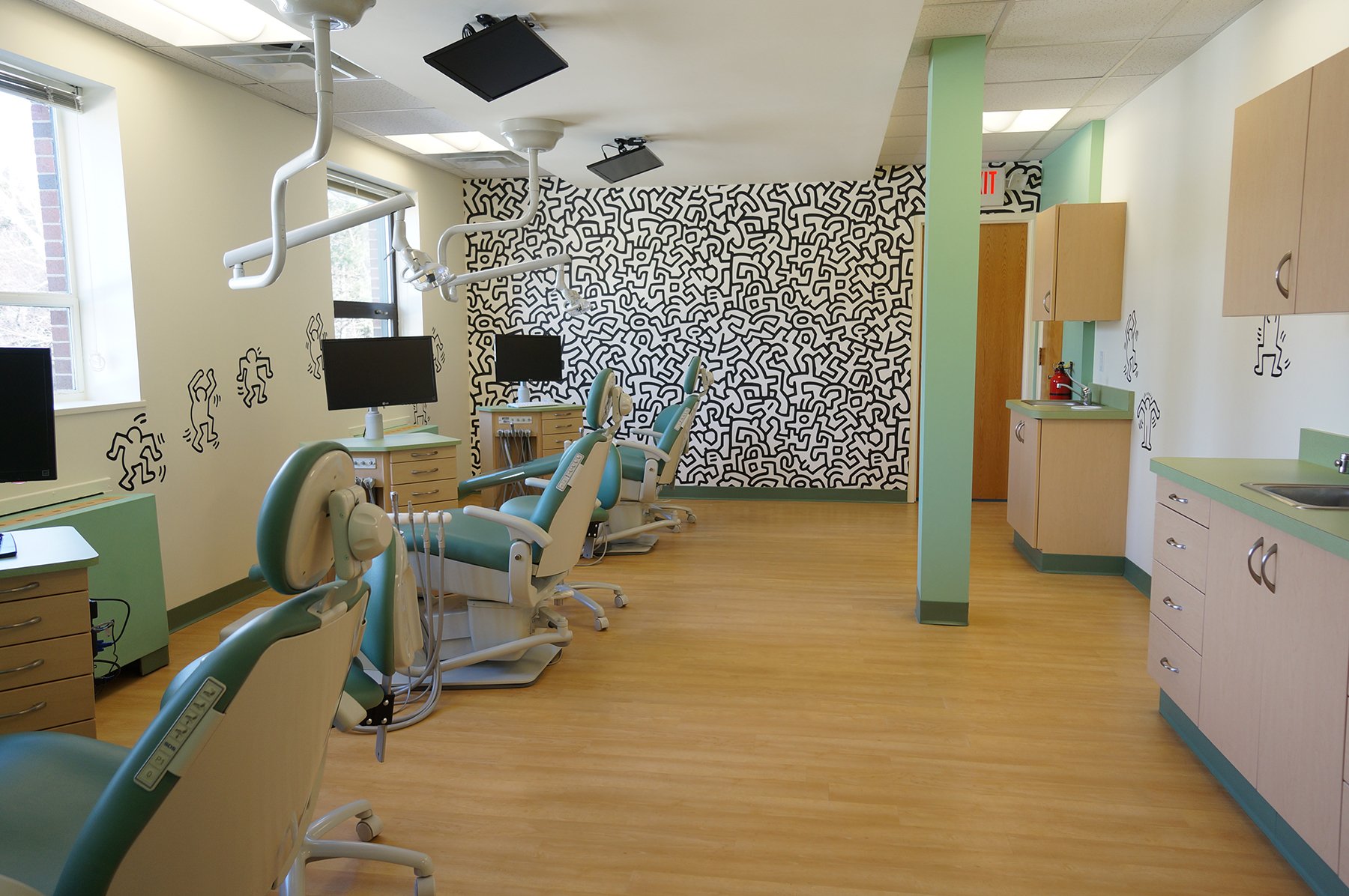 A dental clinic room with dental chairs, monitors, and equipment. The room features a black and white abstract patterned wall, green accents on the chairs and cabinets, and wall decals of dancing figures. There are windows allowing natural light, and ceiling-mounted dental lights and monitors.