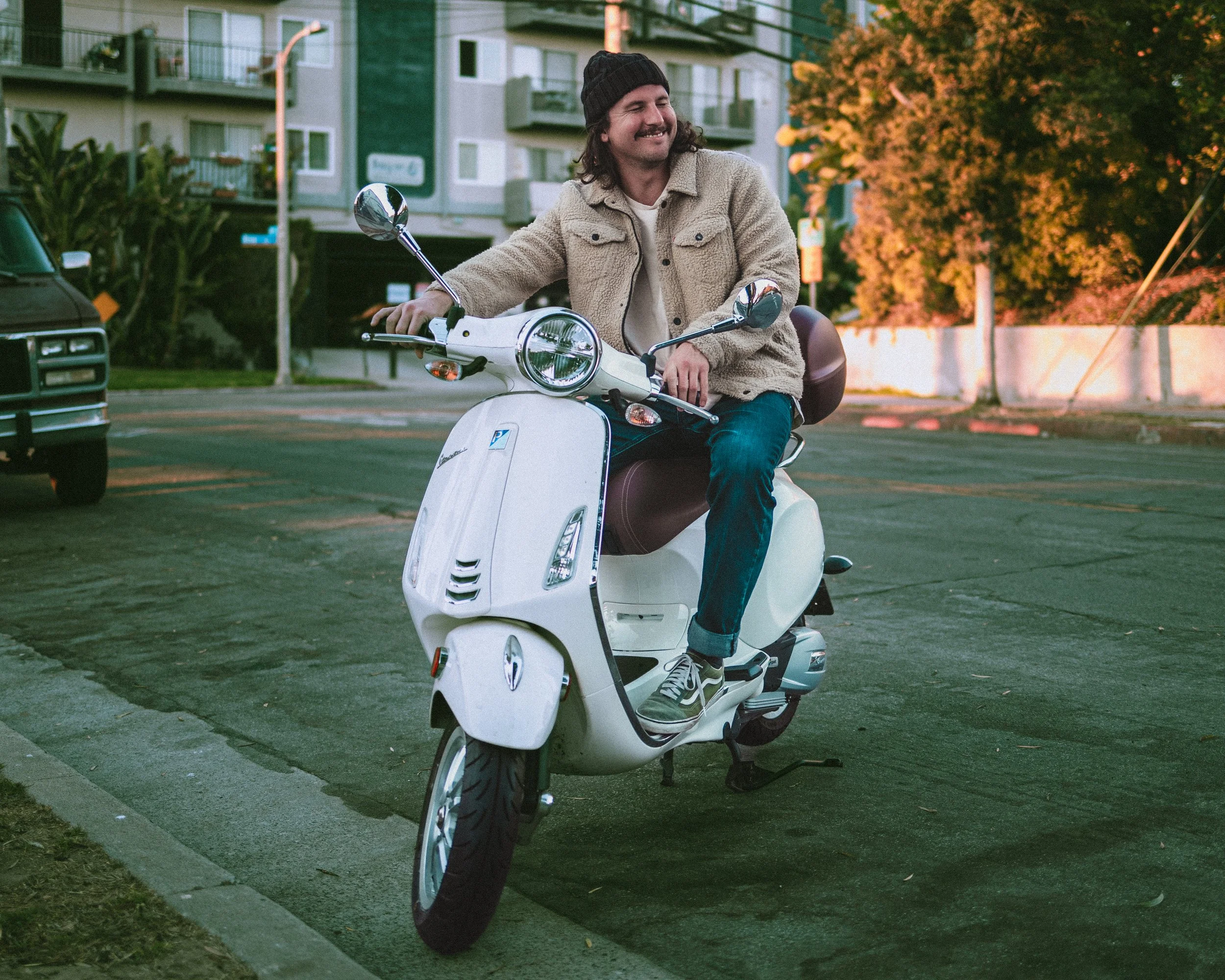 A man with long hair, a beard, wearing a beanie, beige jacket, and jeans, riding a white scooter on a city street during autumn, with trees showing fall foliage in the background.
