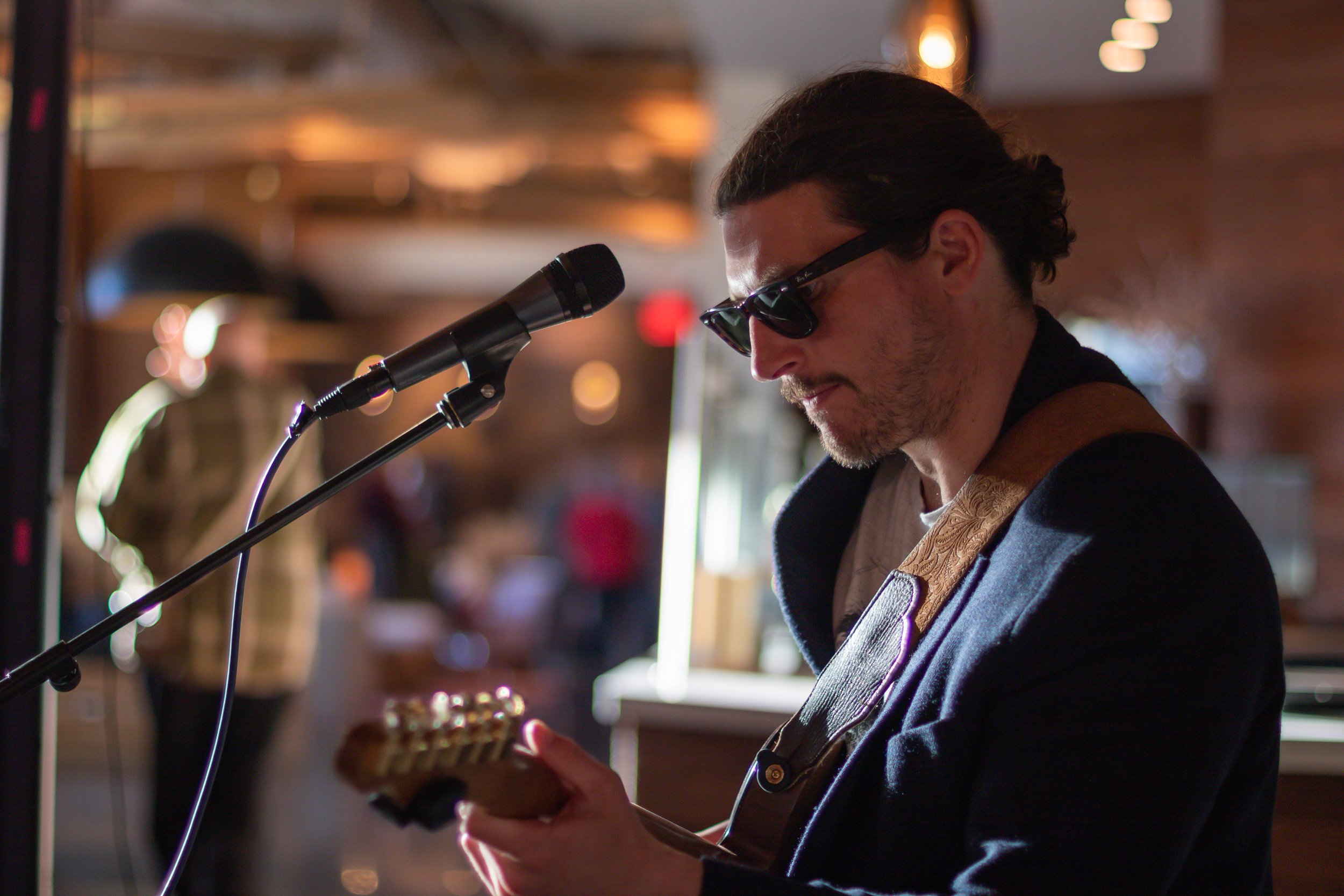 A man wearing sunglasses and a dark jacket playing an electric guitar near a microphone in a warmly lit indoor setting, possibly a cafe or bar.