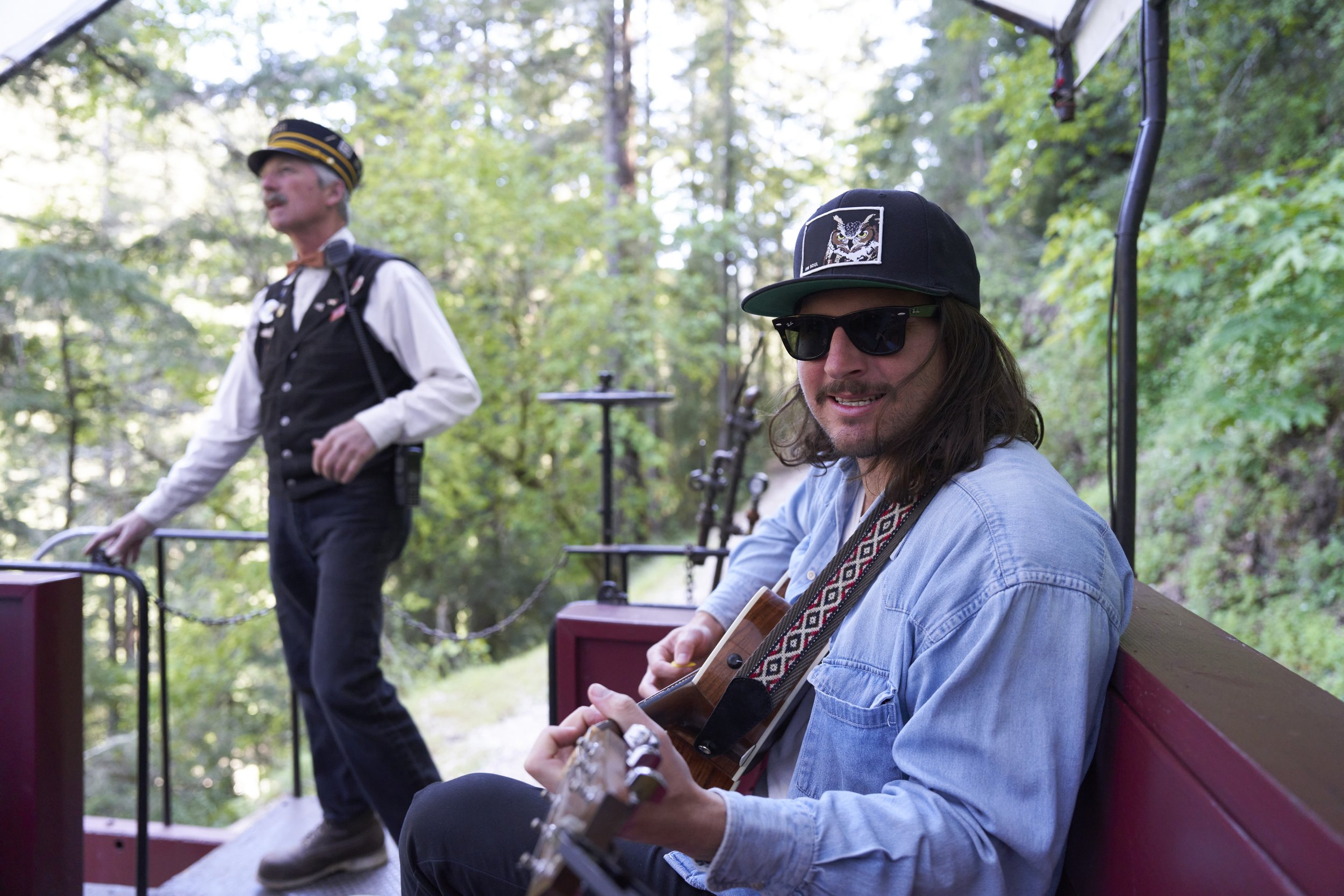 Two men on a park bench outdoors, one playing guitar and the other standing, in a wooded area with trees and sunlight.