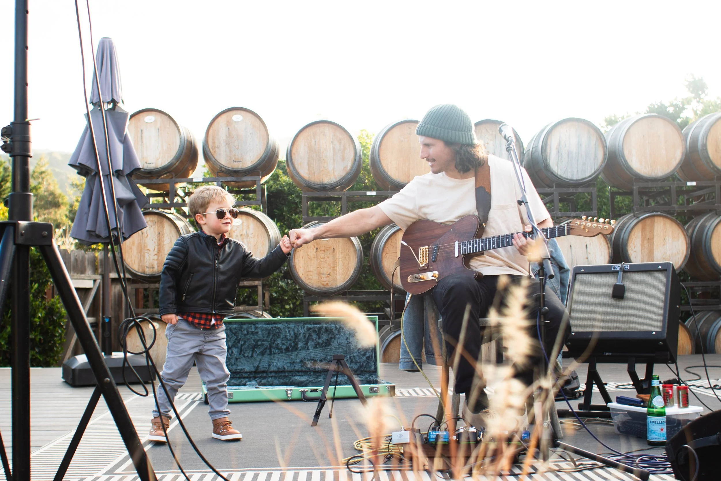 A man playing guitar and a young boy shaking hands on an outdoor stage with wine barrels in the background.