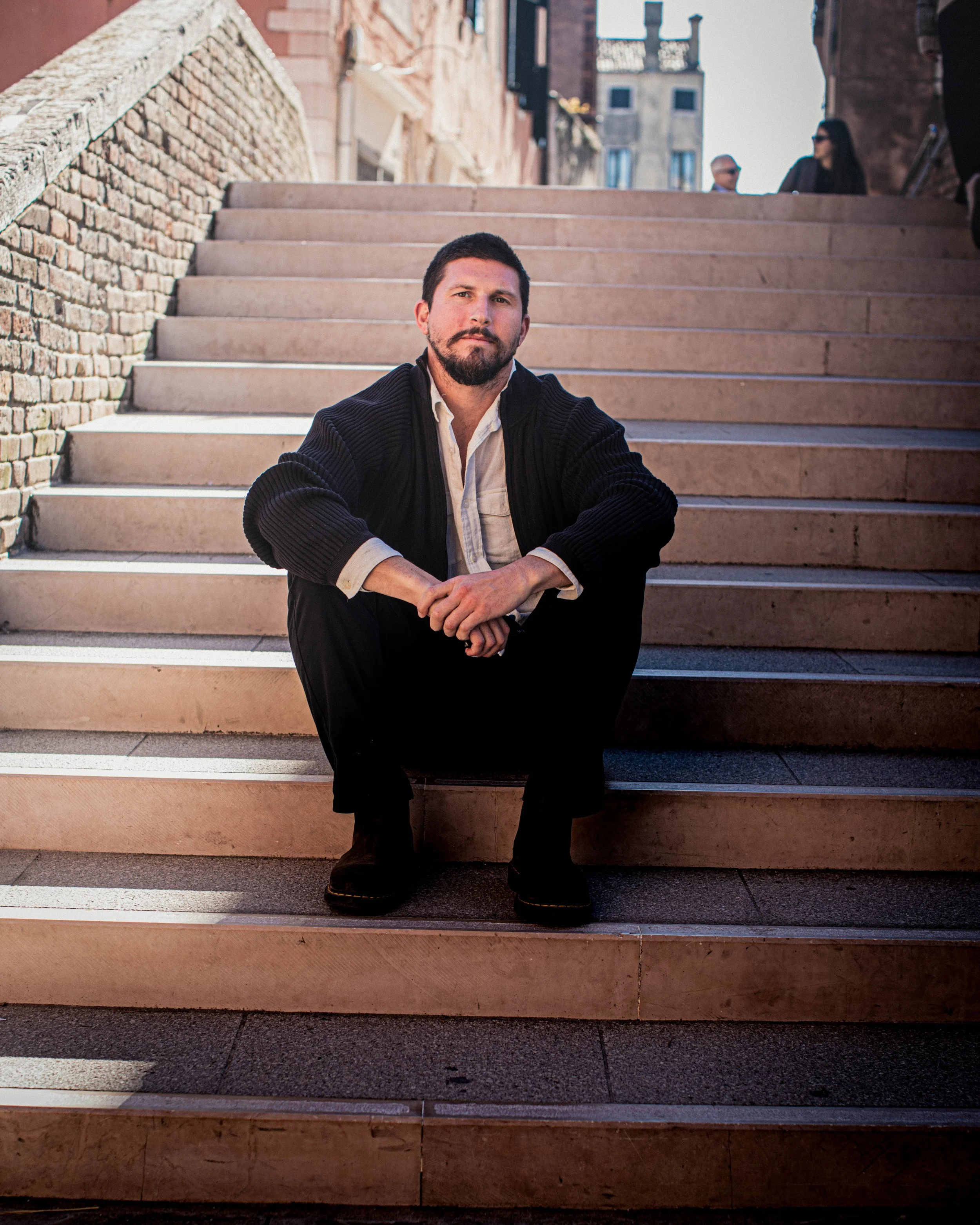 A man with a beard and short hair crouching on a set of stone stairs outdoors, wearing a dark cardigan over a light-colored shirt and dark pants, with buildings in the background.